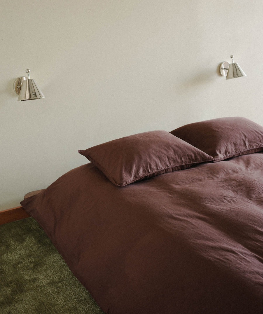 A bed dressed in a dark brown duvet cover and matching pillowcases, photographed in a minimalist bedroom with two silver wall sconces and a light beige wall.