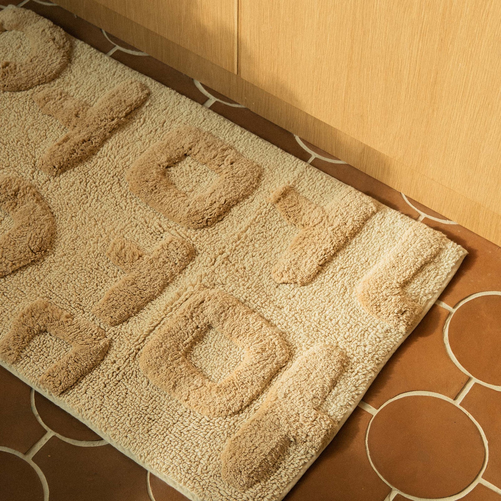 A rectangular beige bath mat with the word 'Mother' written in Korean characters, tufted in varying heights for a textured look on a terracotta tile floor.
