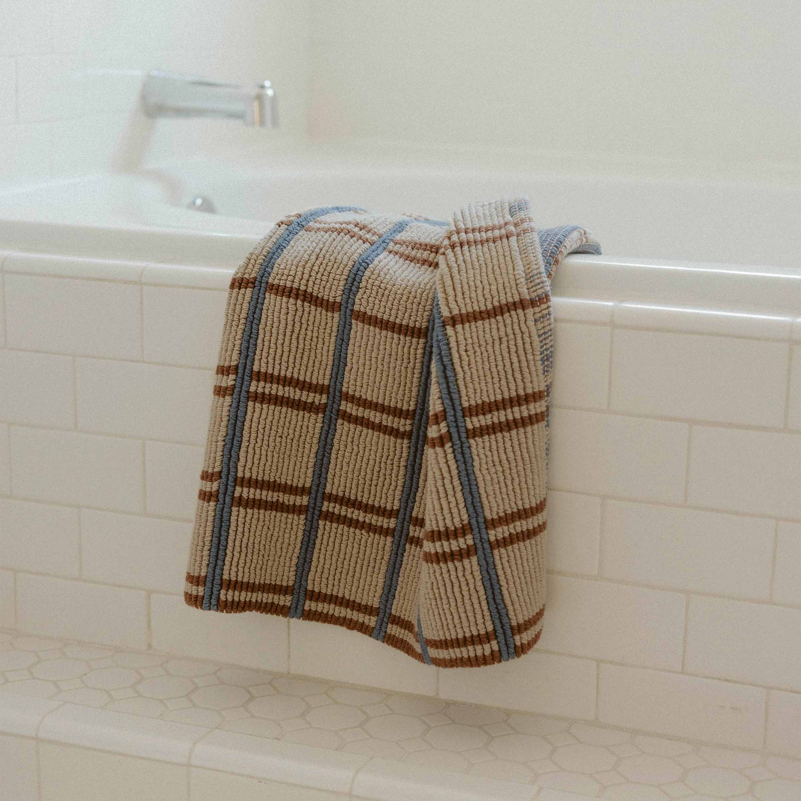 A beige, blue and brown bath mat draped over the side of a white tiled bathtub.