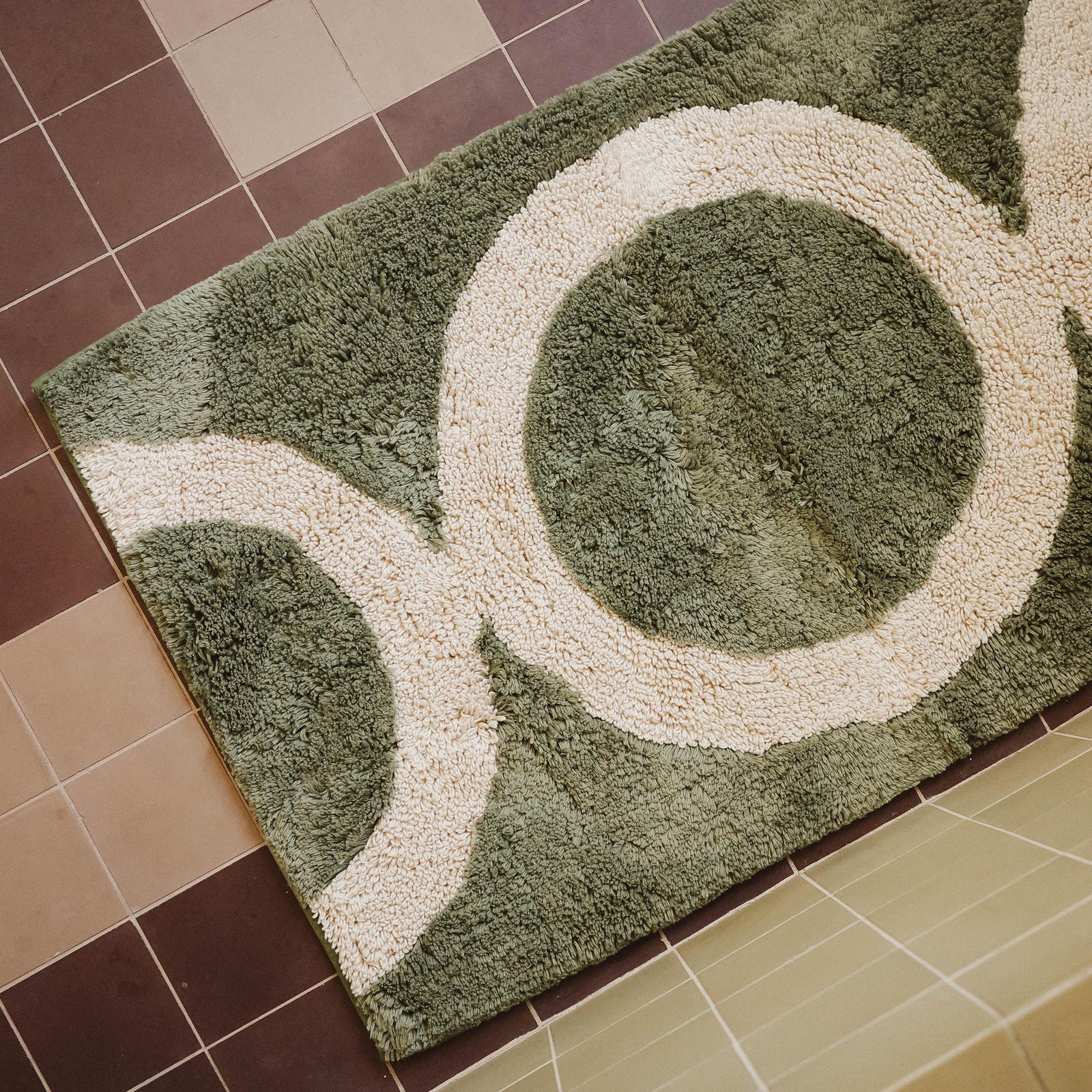 A green tufted bathmat with a beige circle pattern on a tile floor.