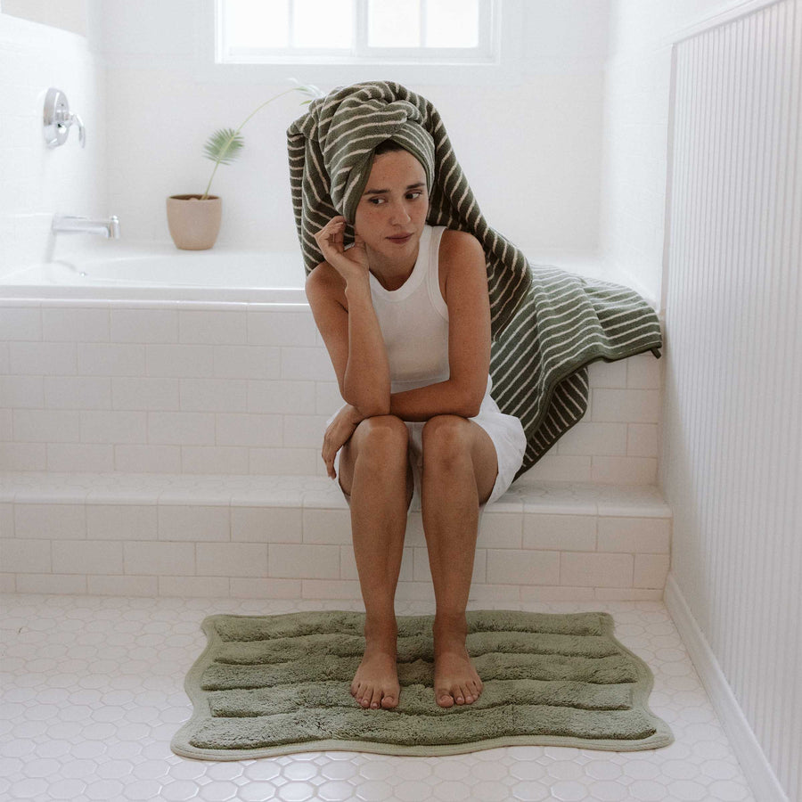 Woman sitting on a green bath mat in a bathroom with a towel draped over her head.