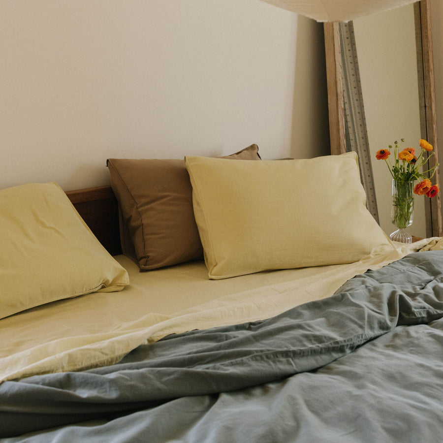 A bed dressed in Límon / light yellow sheets and pillows paired with a Dune / beige pillow and with a green duvet cover in a bedroom setting