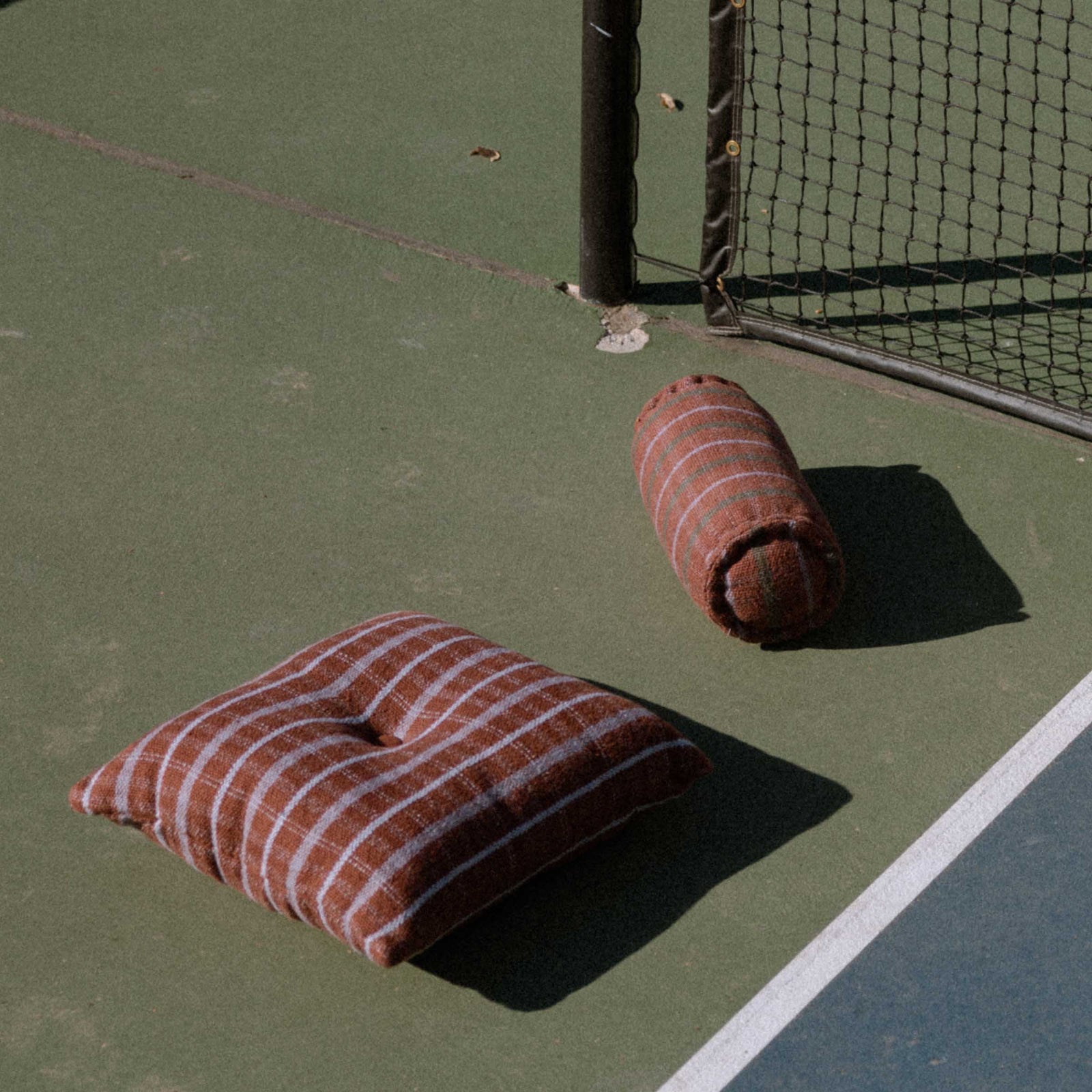 One check pattern brown and lavender throw pillow with a center button and one check pattern brown and lavender bolster on a tennis court.