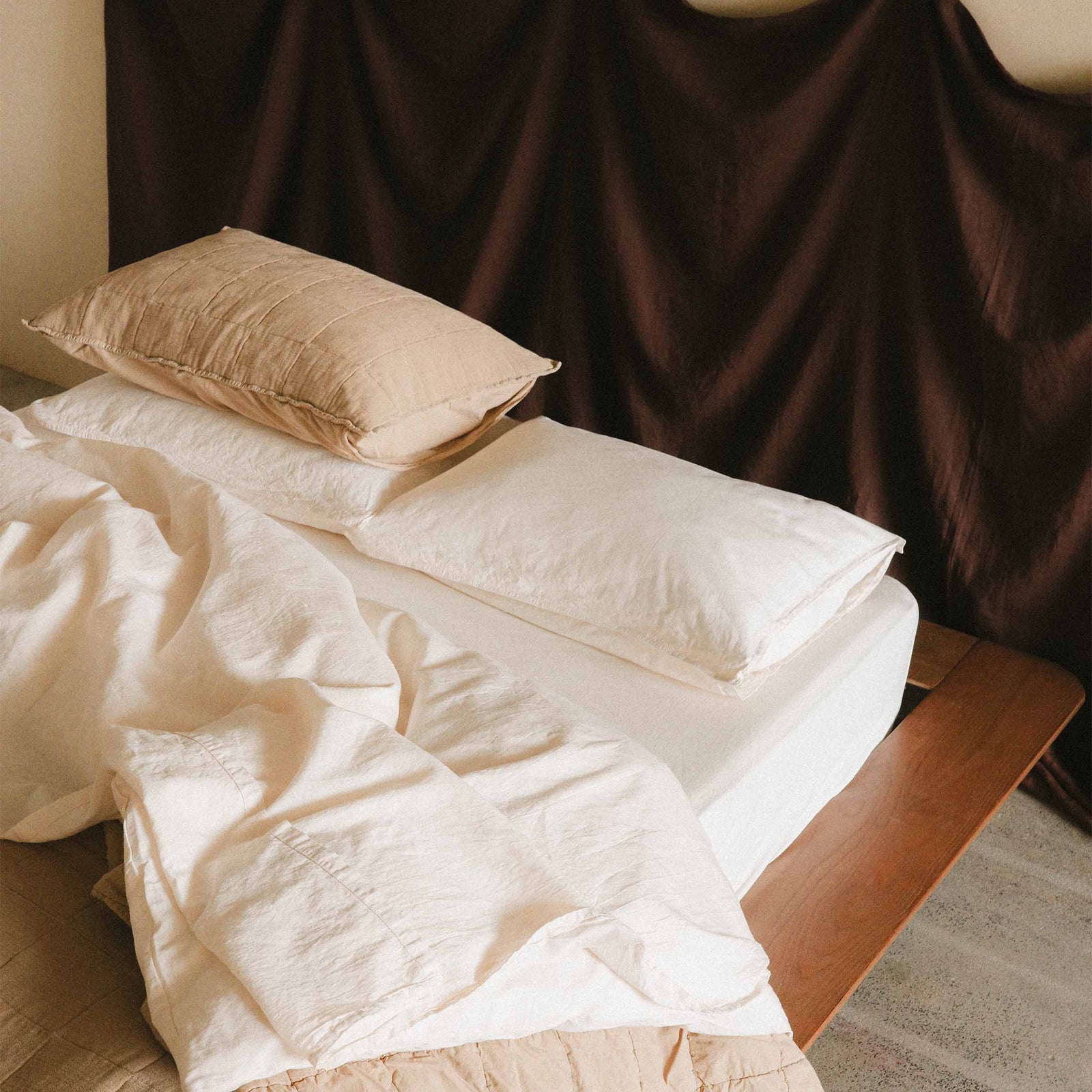 Bed with cream / white sheets and a brown pillow against a purple fabric headboard 