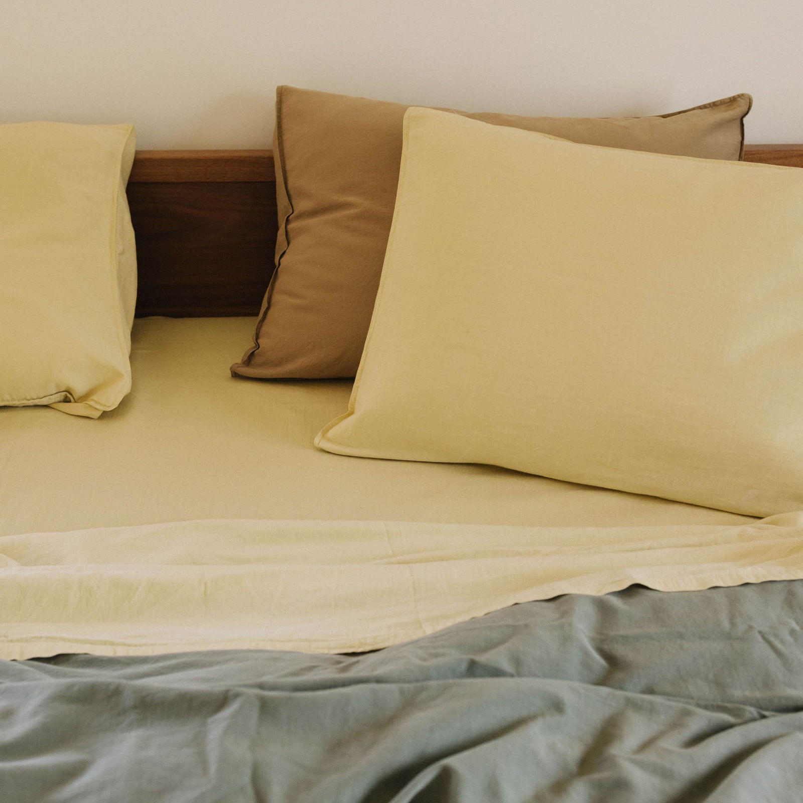 A close up of Límon / light yellow pillows resting against a beige pillow in a bed with a green / blue / Pacific duvet cover