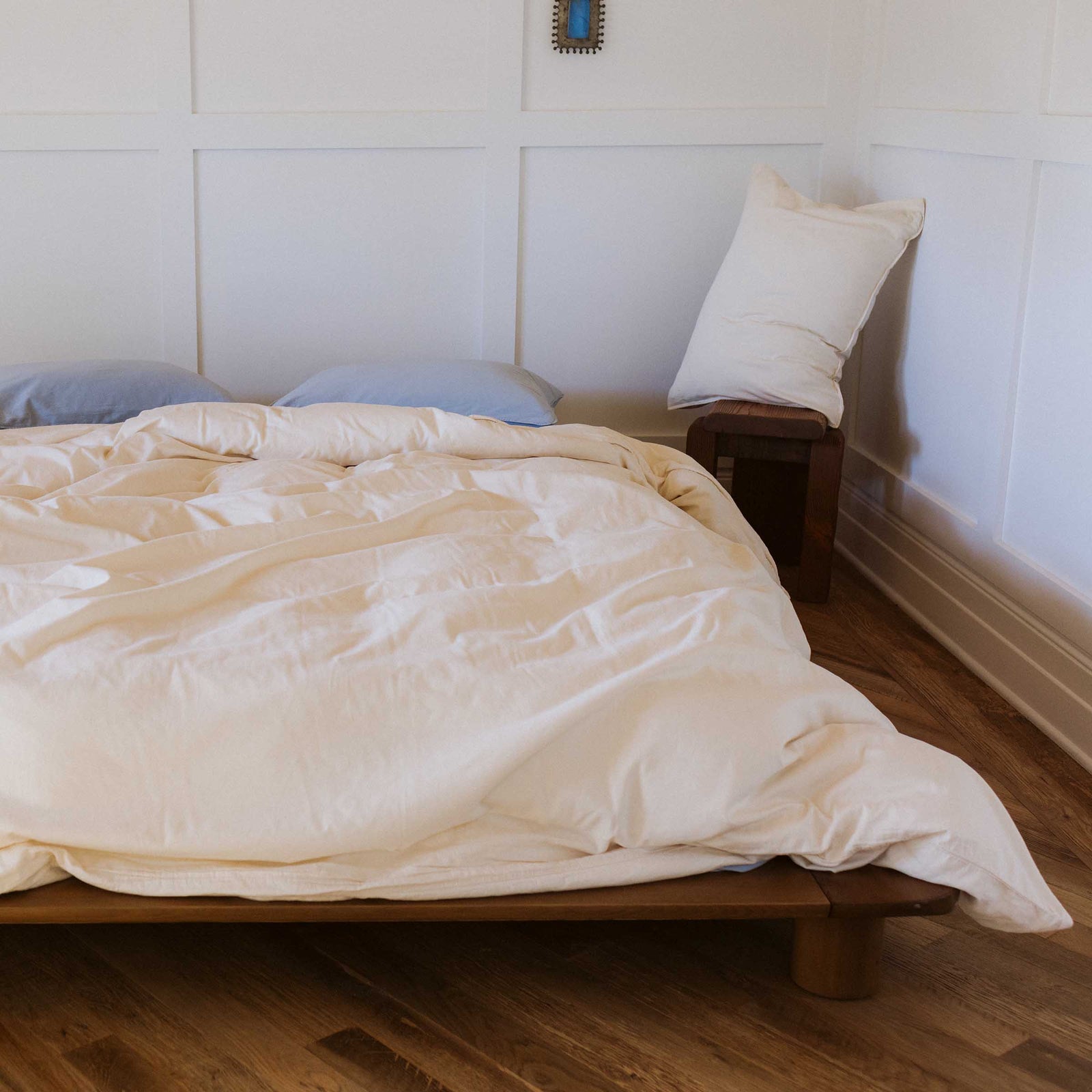 A slightly wrinkled cream colored bed with two blue / Afina pillows in a  bedroom setting