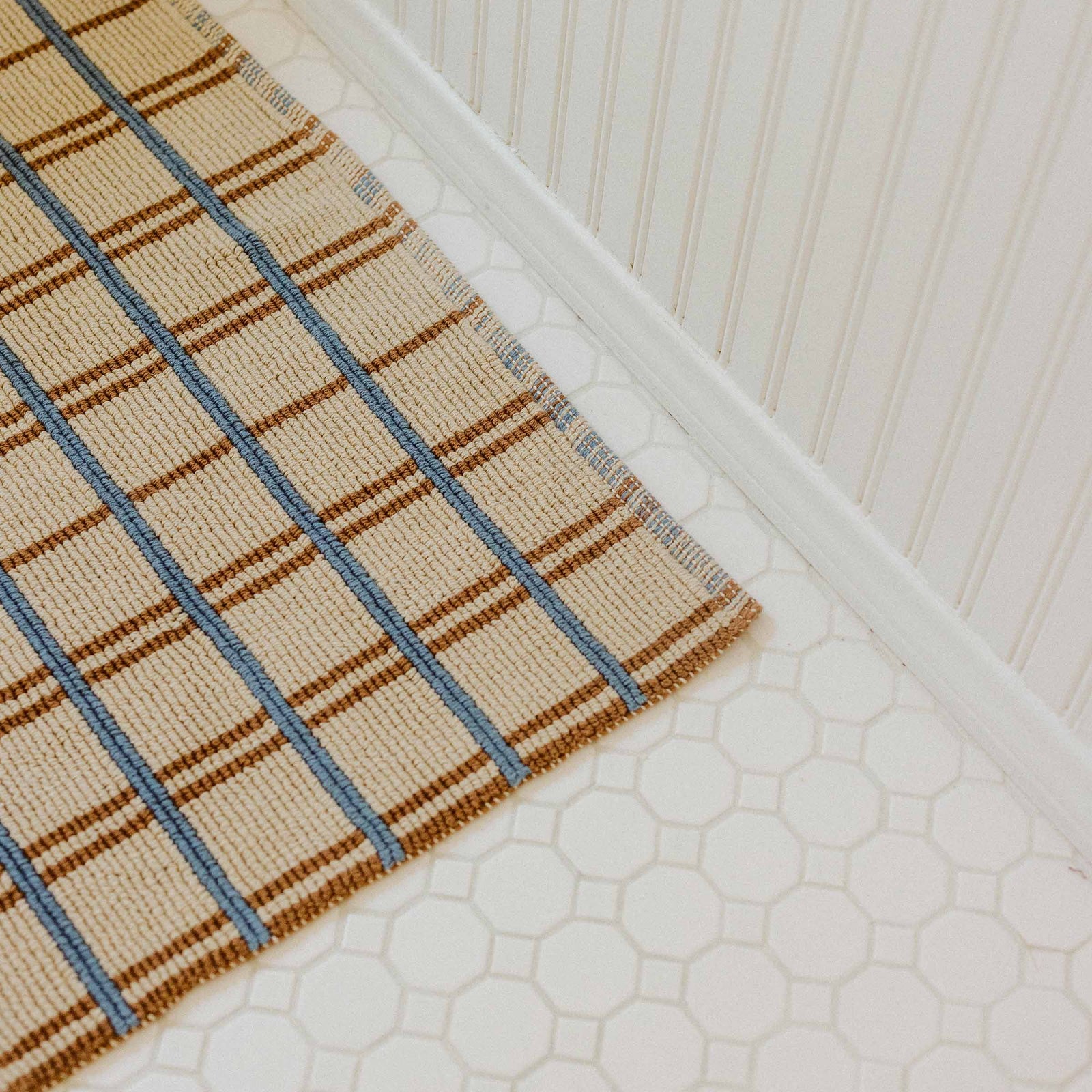 A beige, brown and blue bath mat on a white tile bathroom floor.