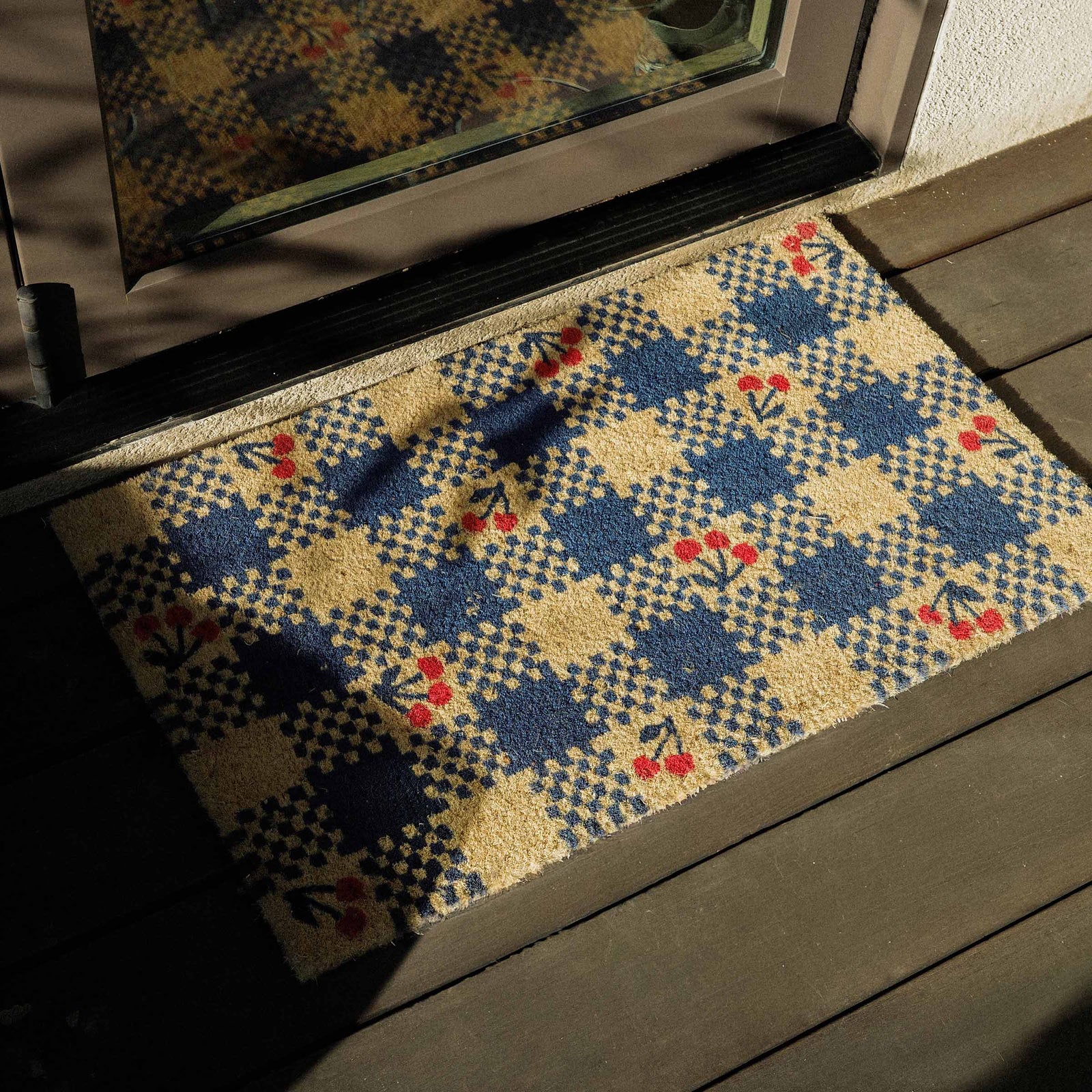 Decorative doormat with a blue geometric pattern and cherries on a wooden deck.