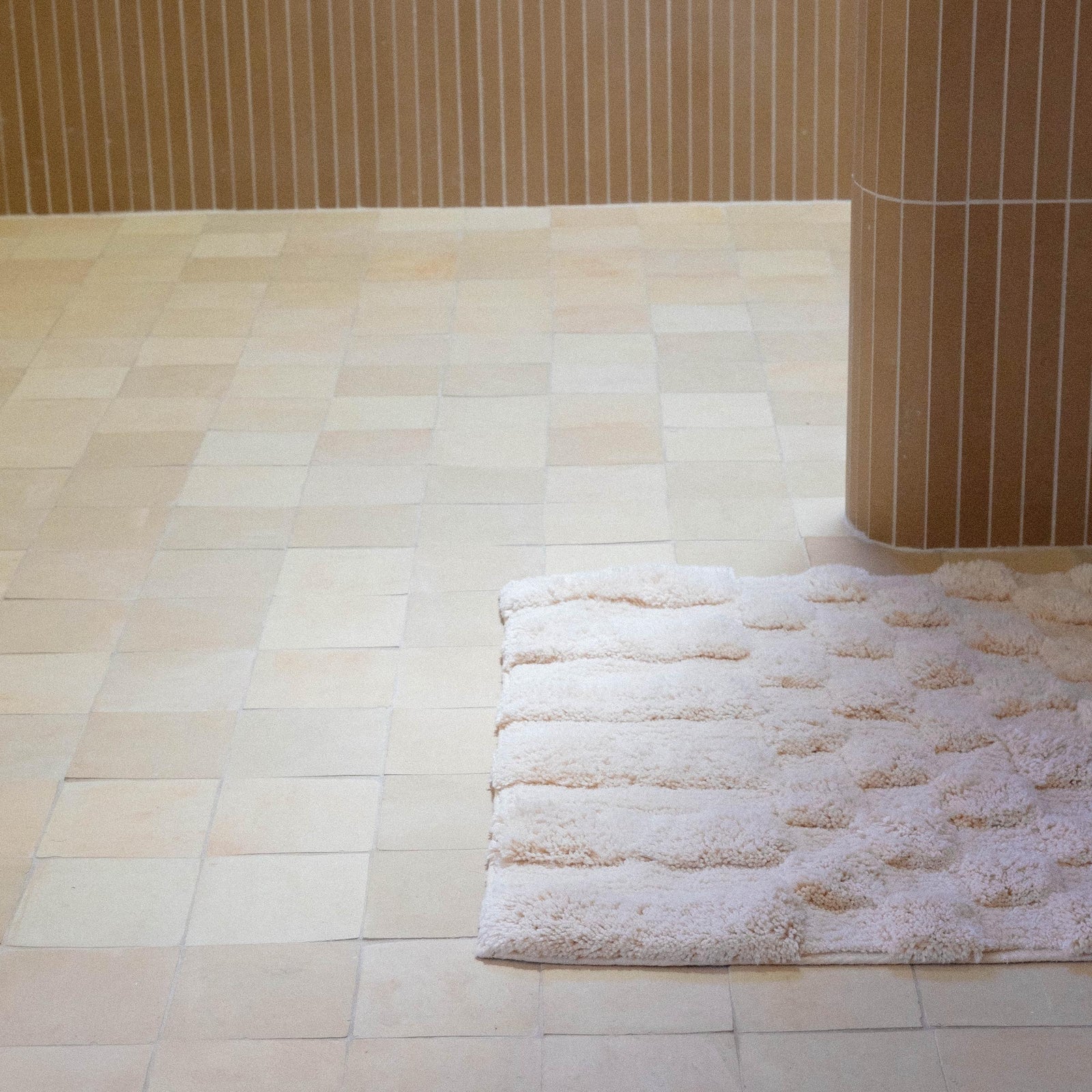 Close up of a pink bath runner with alternating shag heights in a bathroom with brown vertical tiles along the wall.