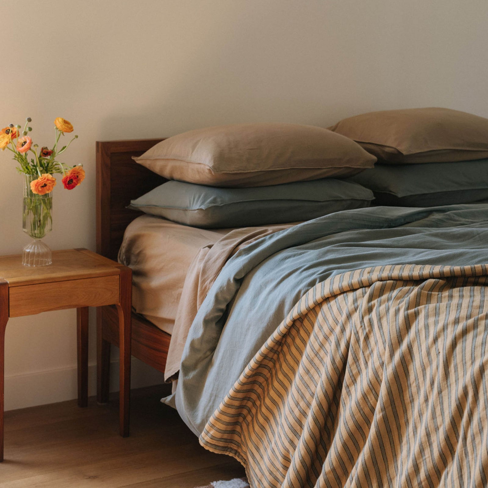 Another angle of a bed in a bright bedroom setting. there is a striped coverlet over a Pacific duvet cover and stacks of pillows in Pacific and Dune over Dune sheets