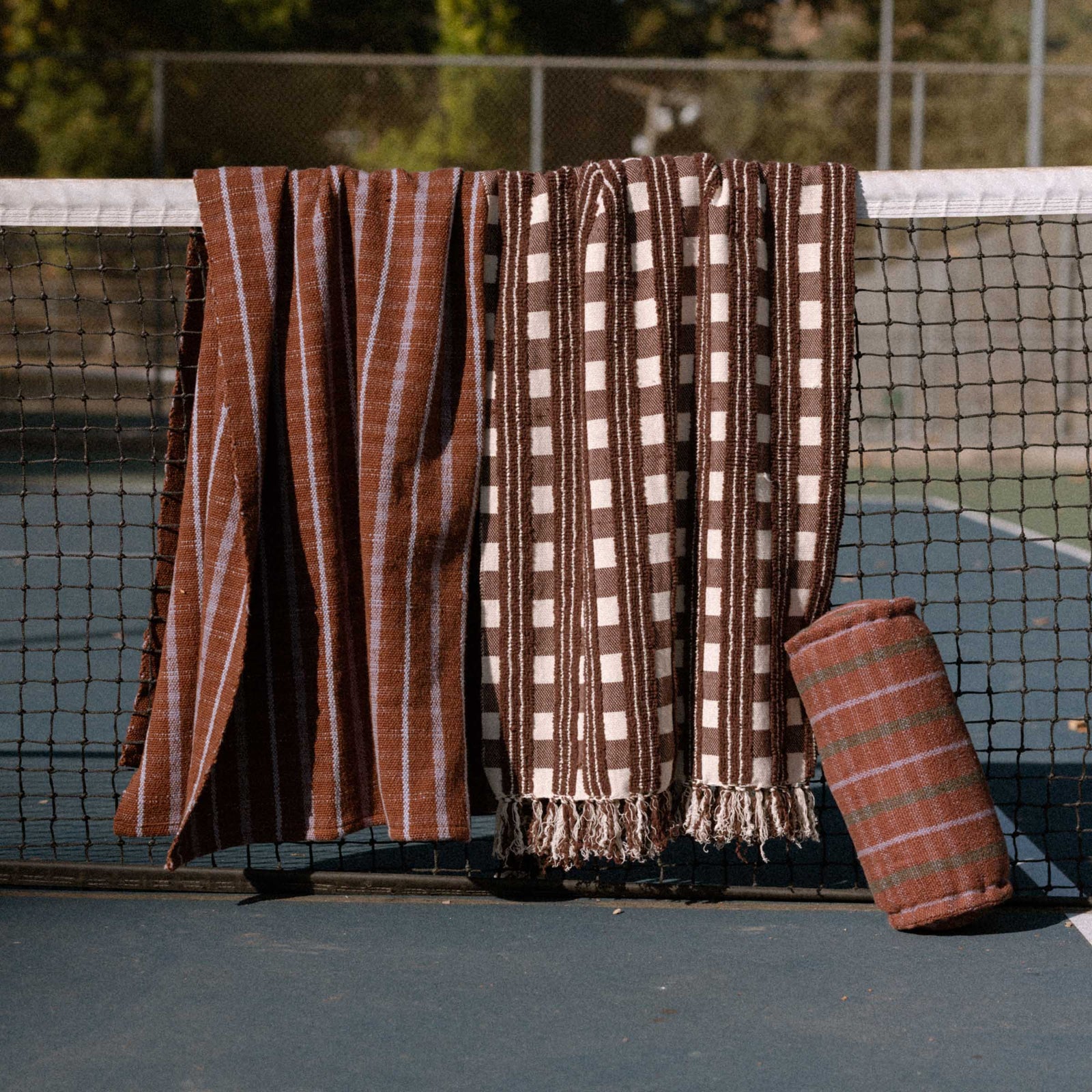 Two brown patterned blankets draped over a tennis net on a court with a brown and lavender bolster leaning against the net.