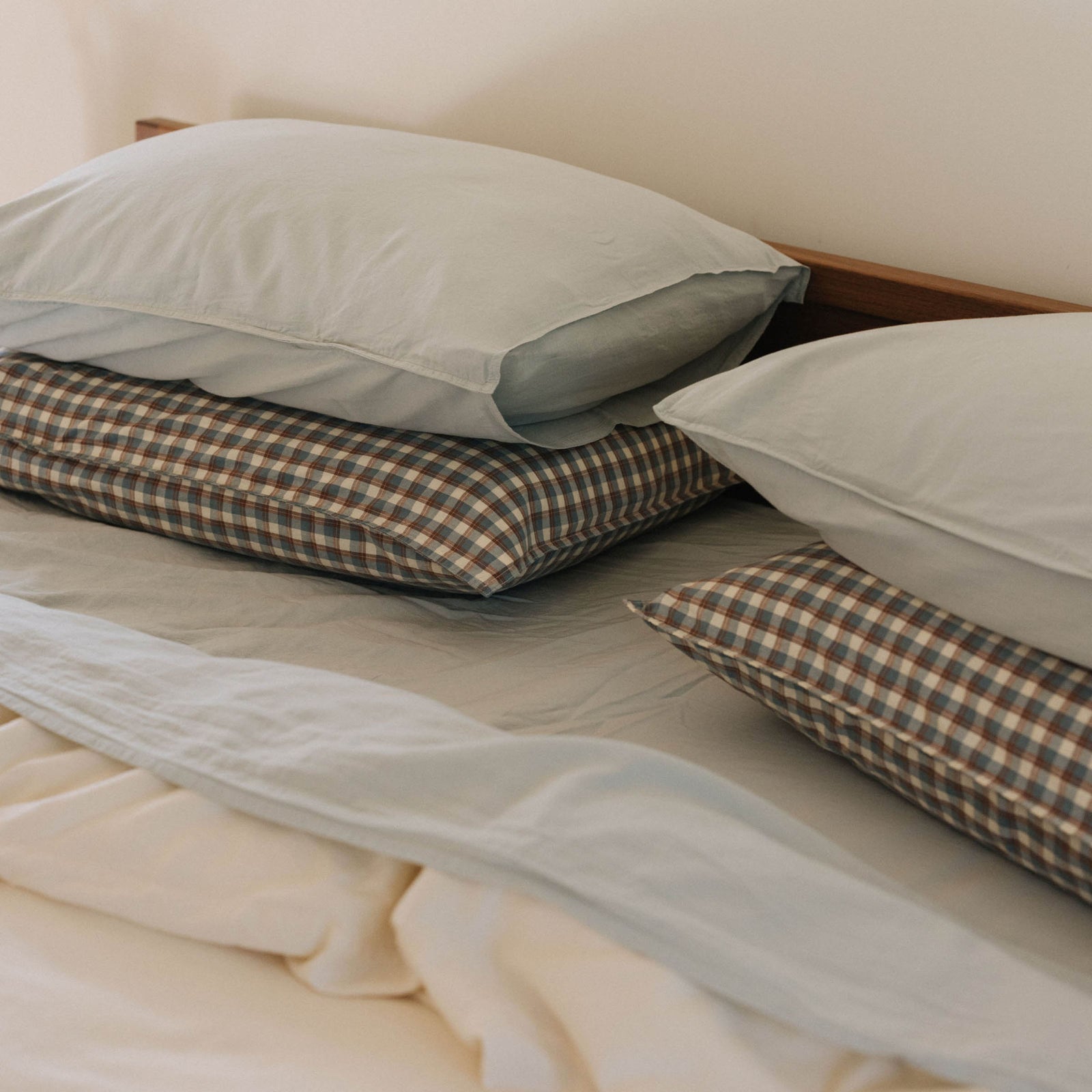 A close up stacked pillows, on the bottom plaid pillows and on top, pale blue / Mist pillows in a bed 