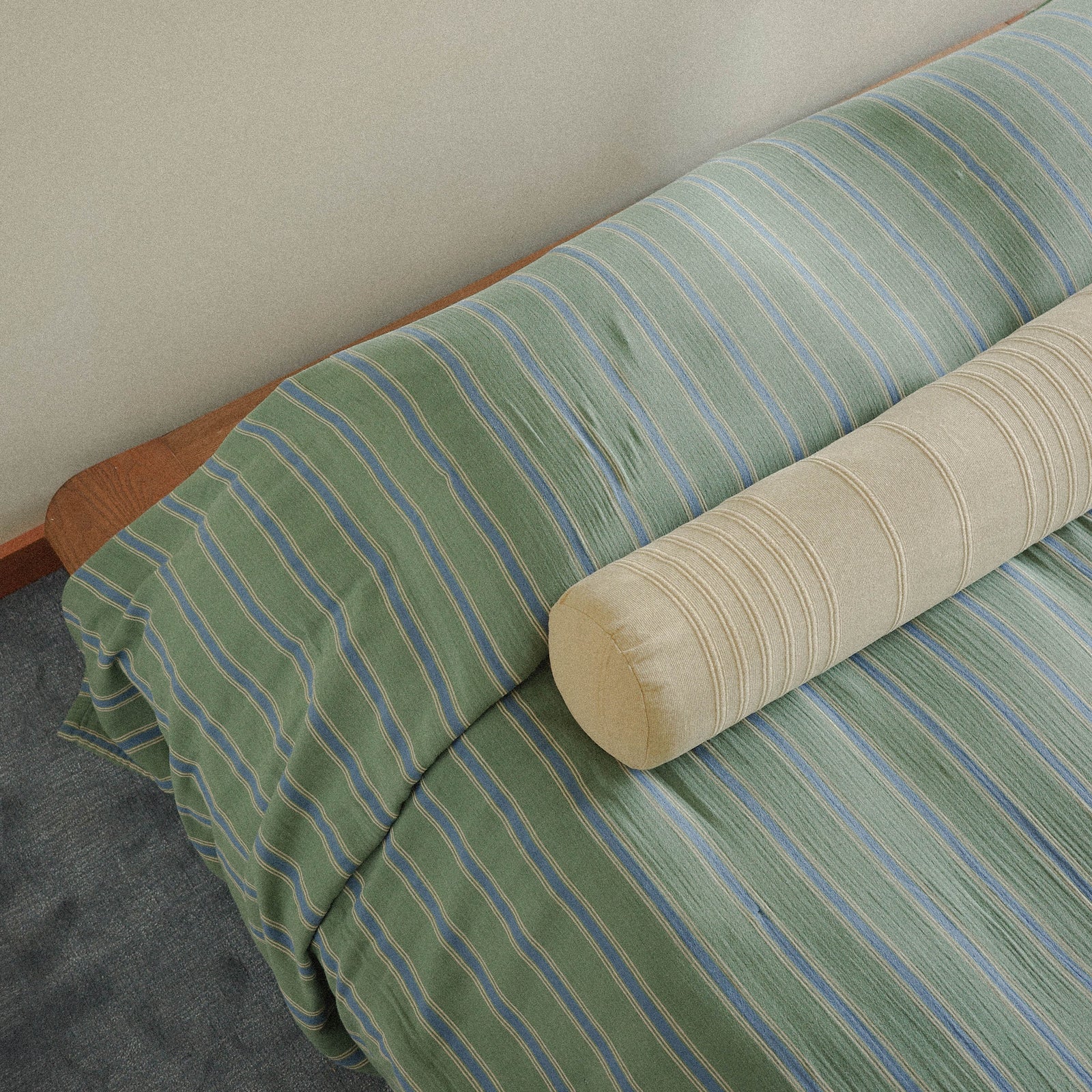 Green and blue striped bedspread with a beige bolster pillow on a bed against a plain wall and green carpet