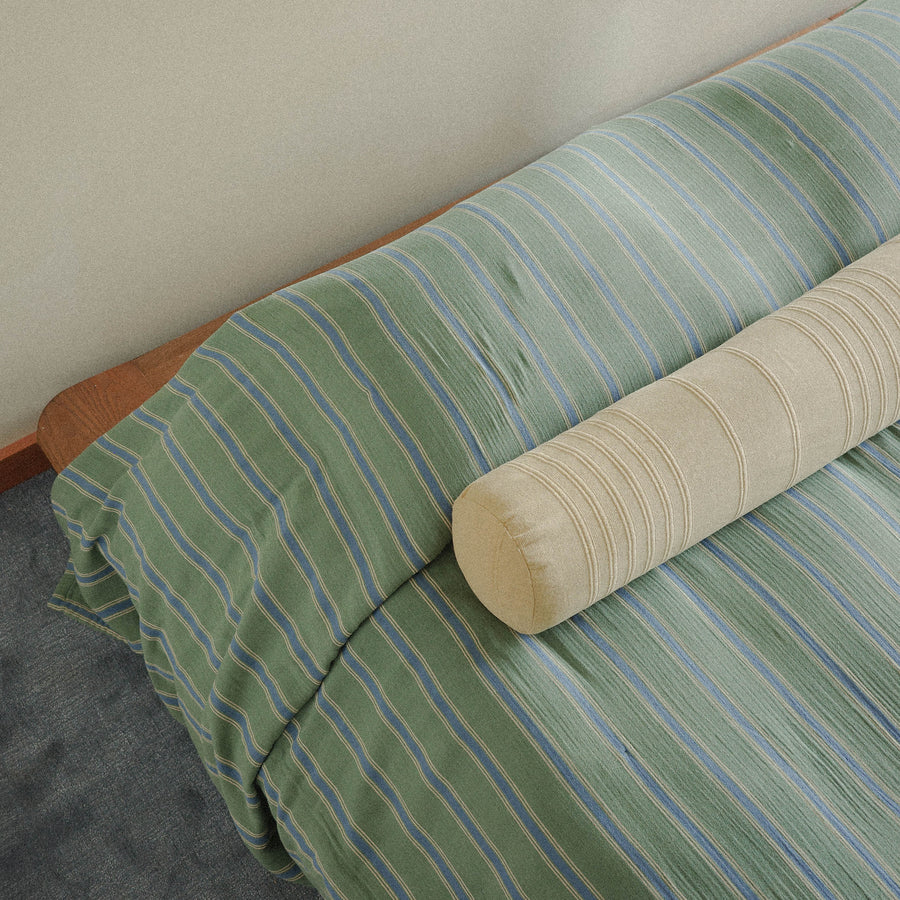 Green and blue striped bedspread with a beige bolster pillow on a bed against a plain wall and green carpet