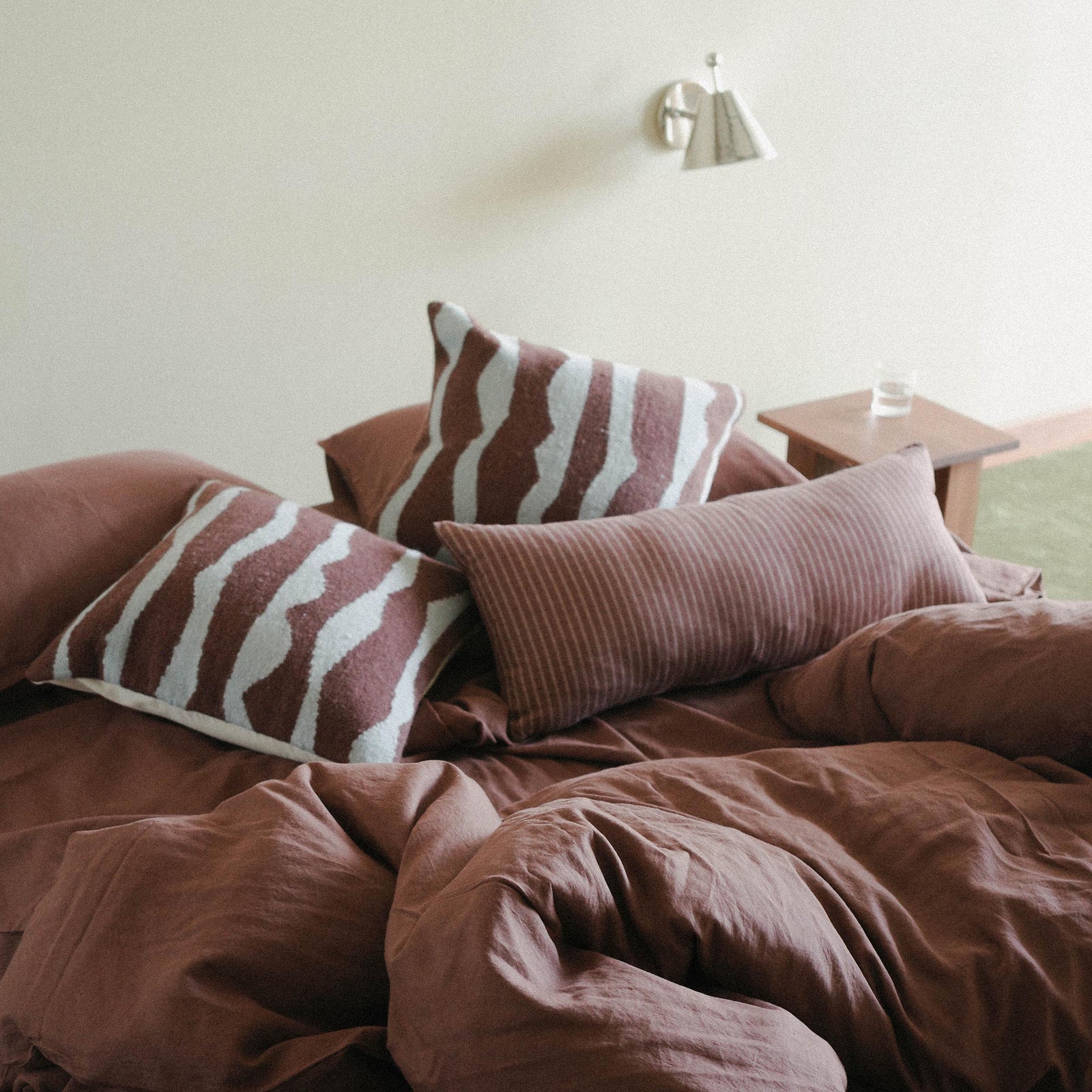 Brown / purple bed with patterned throw pillows and a striped lumbar thrown casually over a duvet cover in a minimalist bedroom