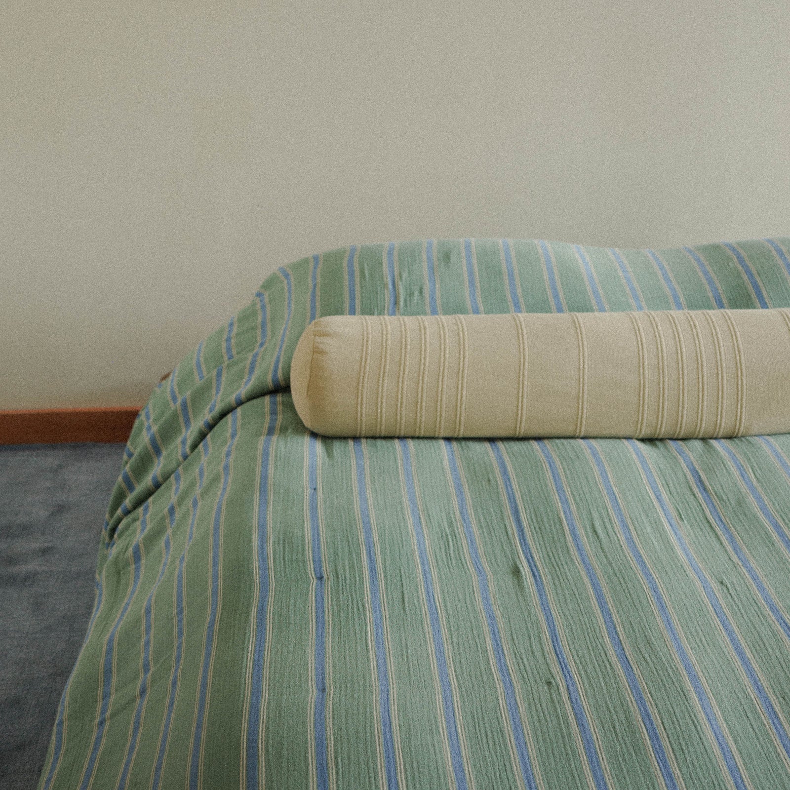 Green striped bedspread with a beige / yellow bolster pillow on a bed against a plain wall