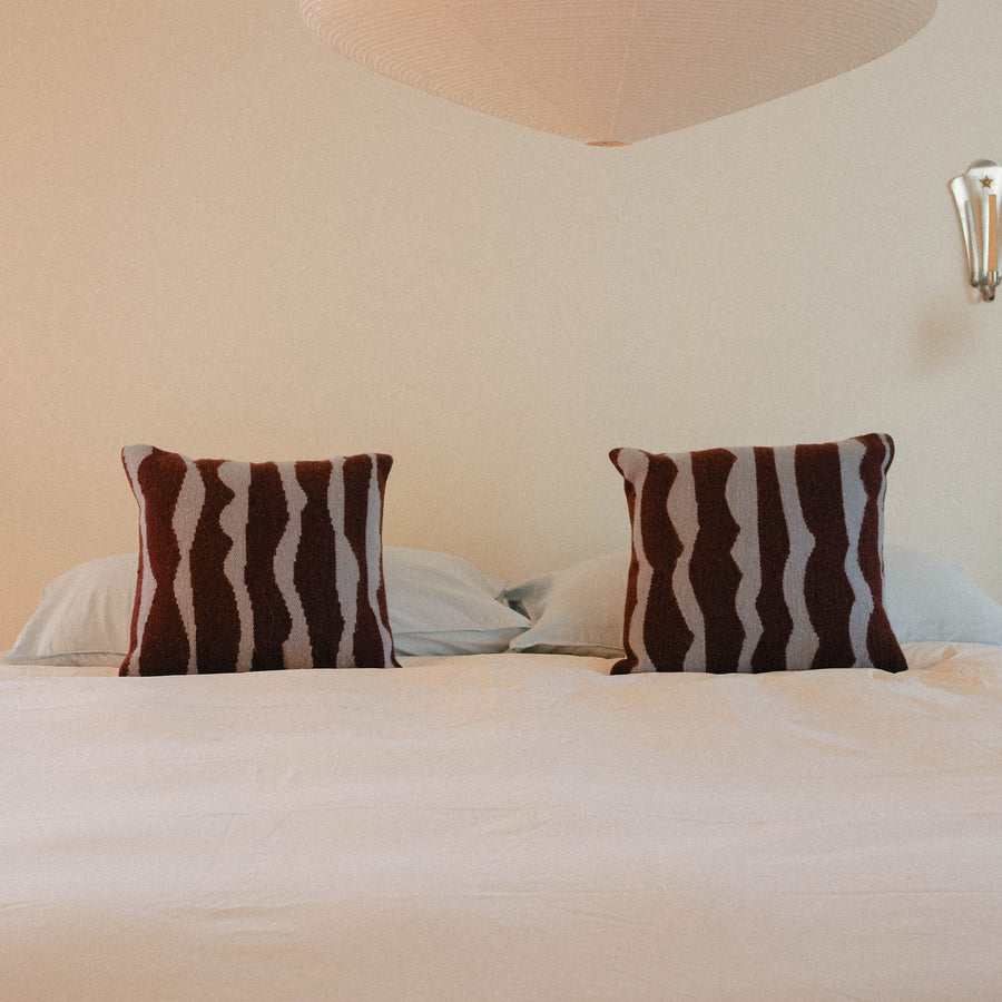 A bed dressed in light blue sheets, cream duvet with two brown and blue organic striped pillows, positioned in a warmly lit bedroom.