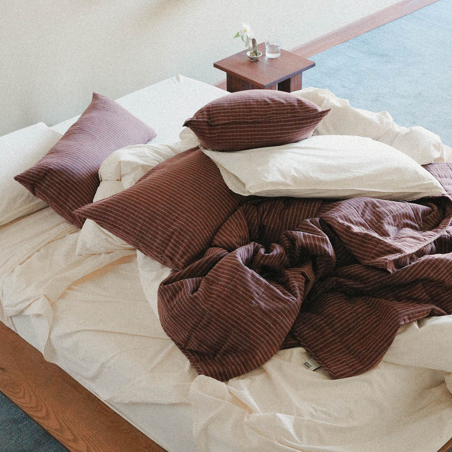 A messy bed with cream sheets, pillows and a striped duvet cover and lumbar pillow in a bright bedroom