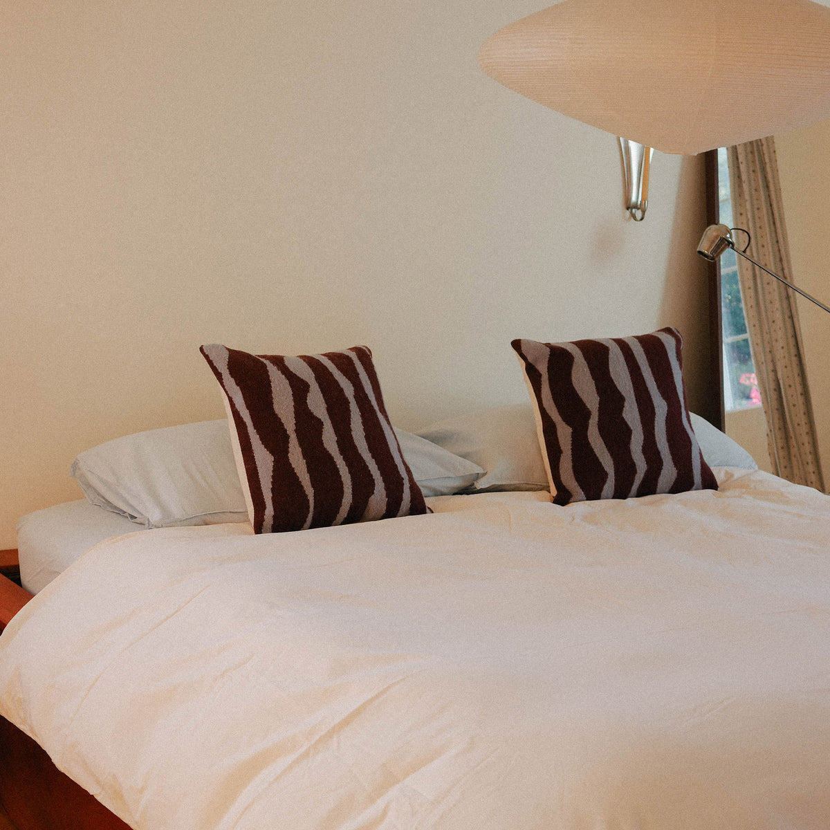 A bed dressed in light blue sheets, cream duvet with two brown and blue organic striped pillows, positioned in a warmly lit bedroom.