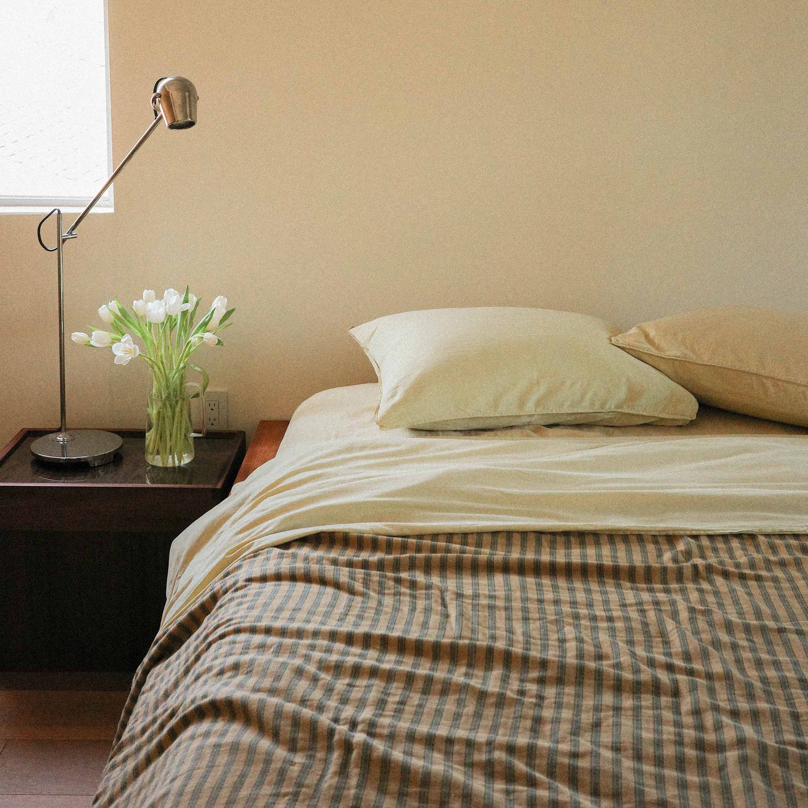 Bed with yellow sheets and a striped coverlet next to a nightstand, and lamp near a window.