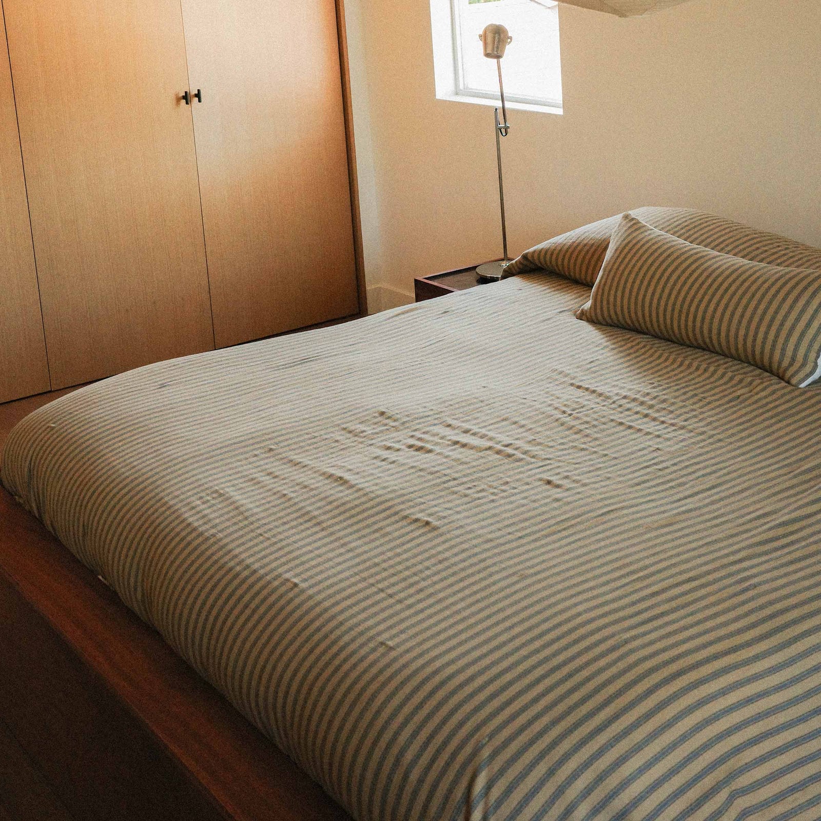 Bed with striped coverlet pulled over two pillows with a lumbar in front in the same pattern in a room with wooden wardrobe and lamp.