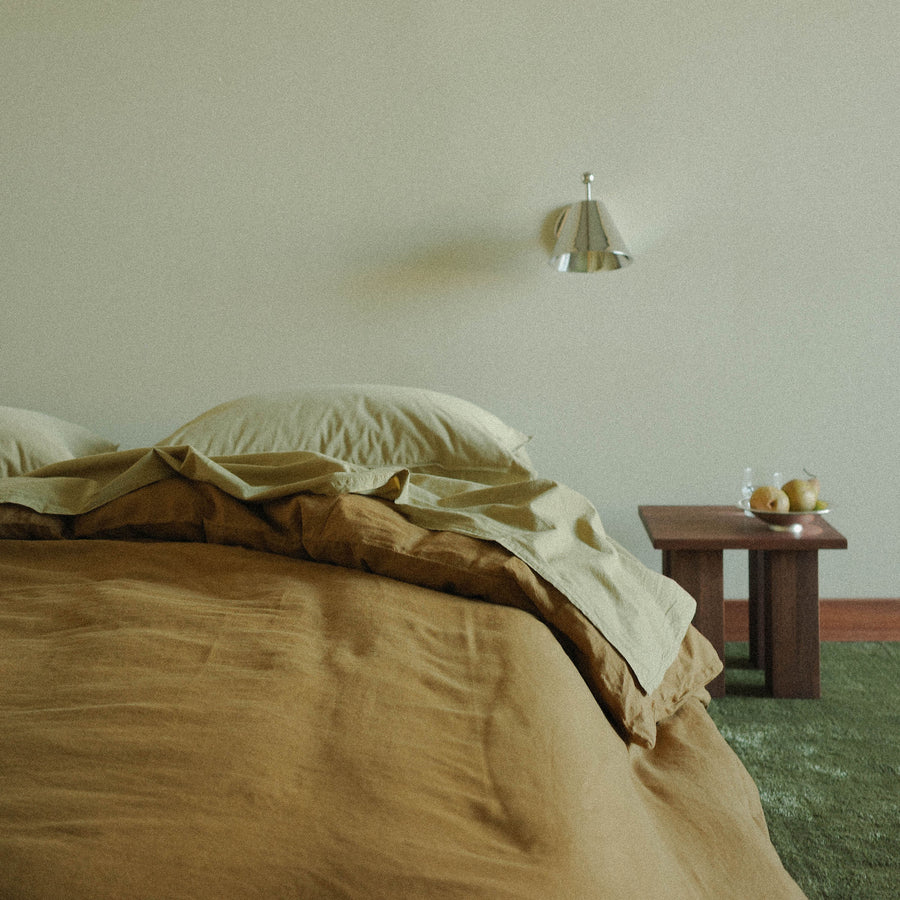 Bed with brown and beige bedding in a room with a wall sconce and small nightstand with a bowl of fruit .