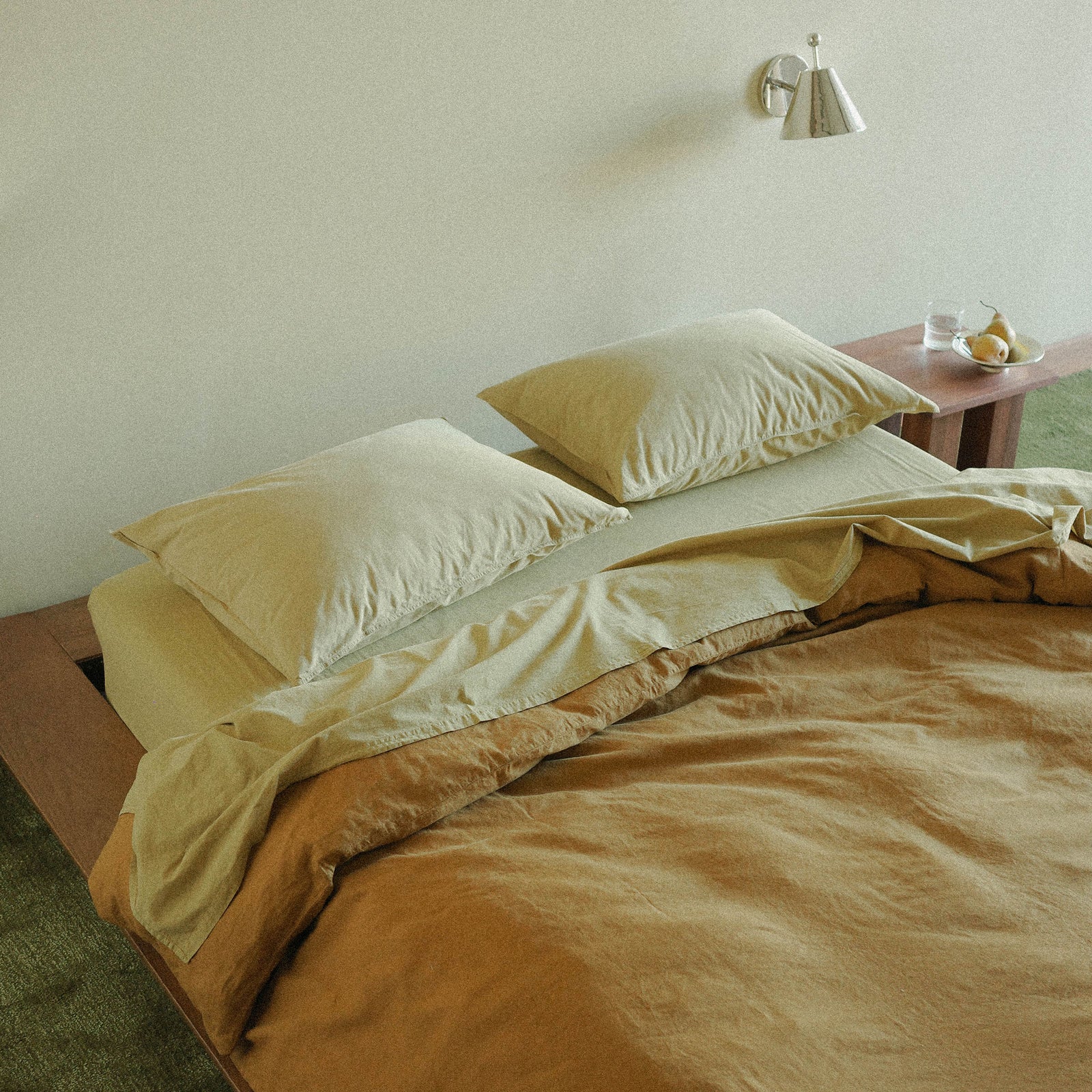 Bed with beige and brown bedding and pillows in a room with a chrome wall sconce and small nightstand.