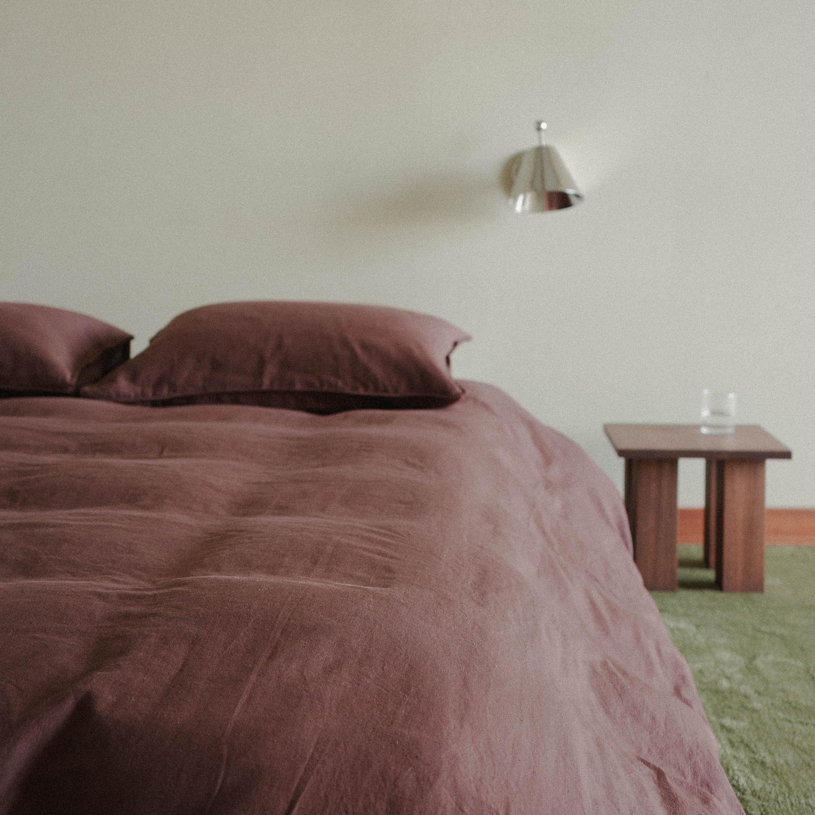 A closer photo of the bed with a brown / purple duvet cover, the room is tastefully sparse with a green carpet and a wood nightstand 