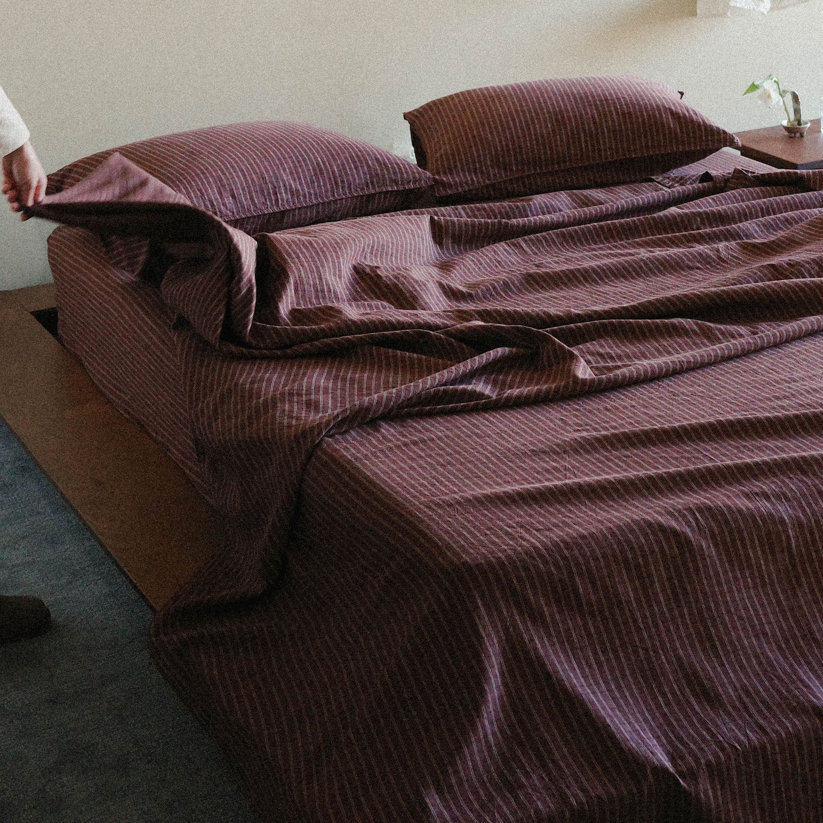 Brown striped bedspread on a bed with pillows in a bedroom setting with a hand pulling a corner of the sheet