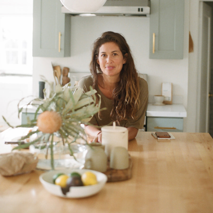 A woman with long brown hair sits at a light wood table in a bright, modern kitchen with sage green cabinets. In the foreground, a white bowl with lemons and a decorative plant arrangement are visible on the table.