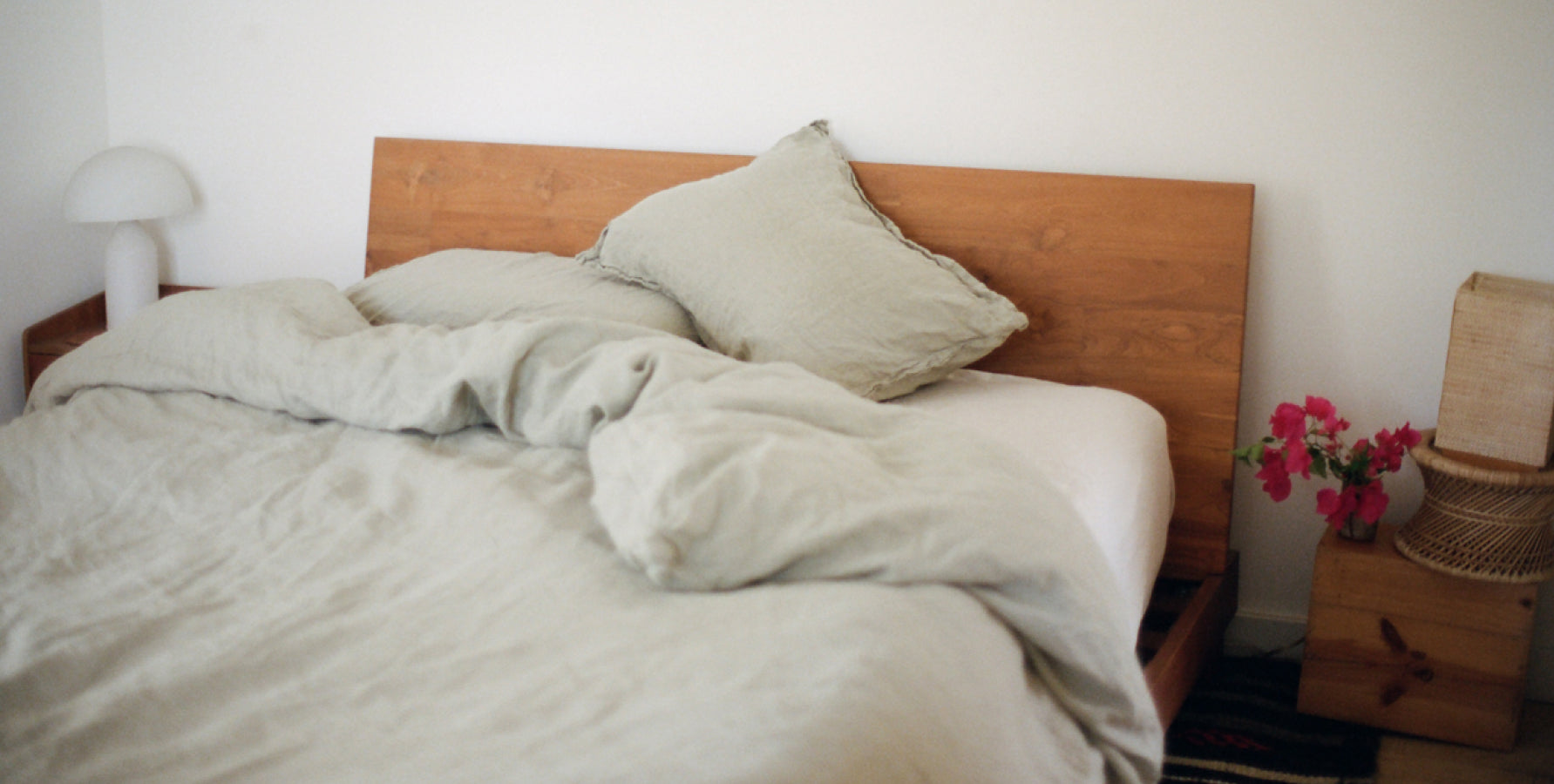 A messily made bed with sage green linen bedding, a wooden headboard in a bright, minimalist bedroom.