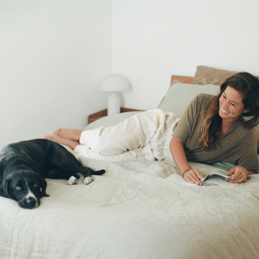 A woman lies on a bed reading a book, smiling, with a black dog resting on the bed beside her. The room has white walls and bedding, with a small white table lamp in the background.