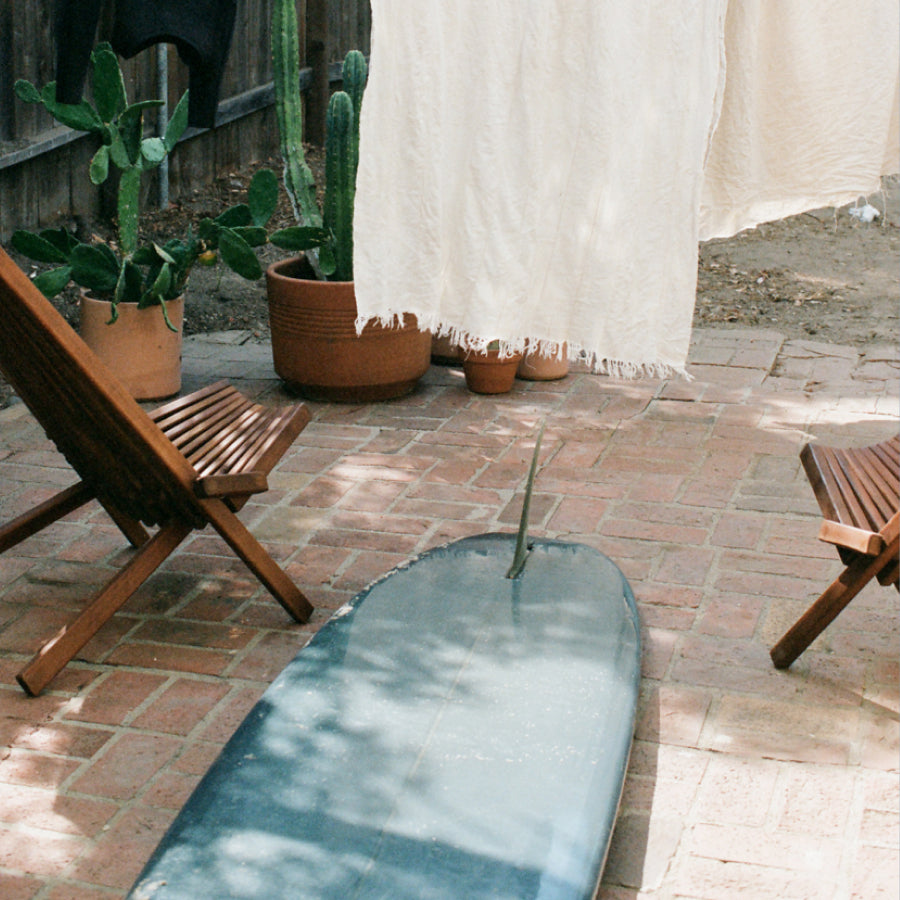 A dark teal surfboard lies on a brick patio in an outdoor setting, surrounded by wooden lounge chairs, potted cacti, and a cream-colored textile hanging in the background.