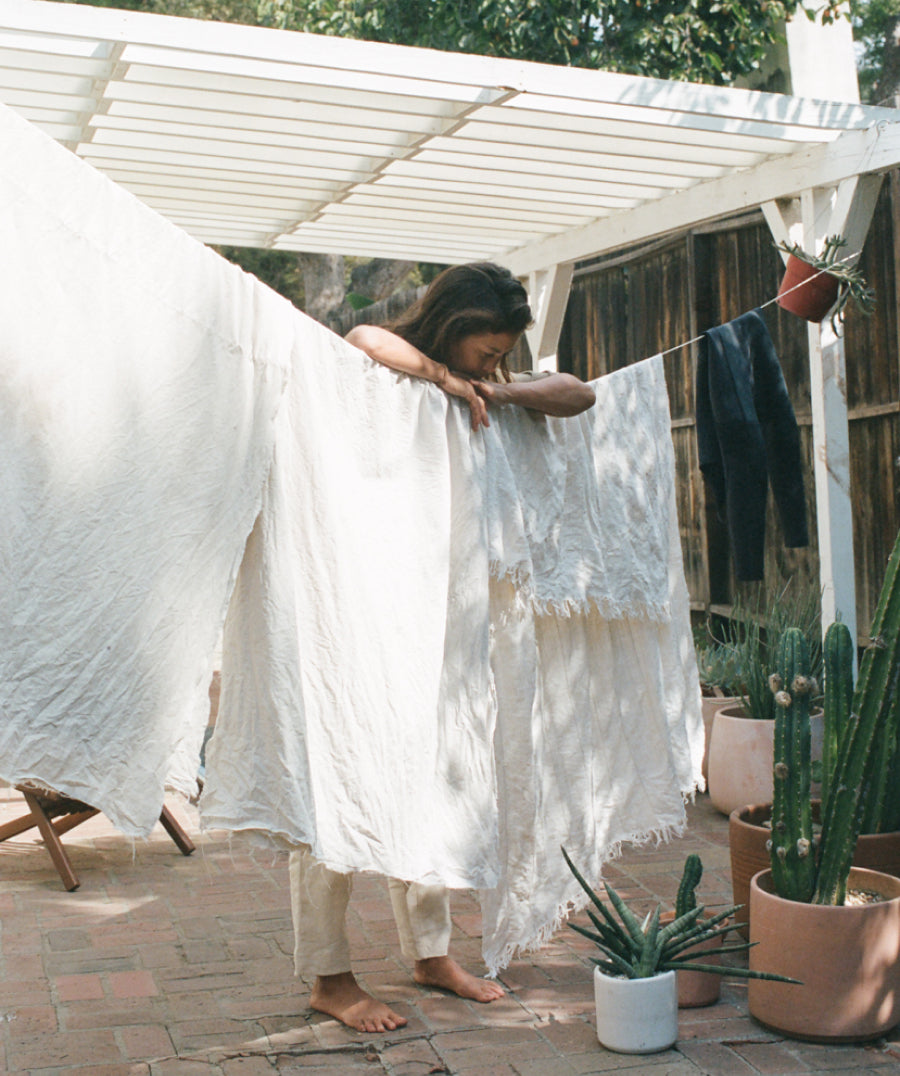 A woman stands barefoot under a white pergola outdoors, holding up large white textured fabric sheets or linens on a line. The scene includes potted cacti, a wooden fence, and a brick patio.