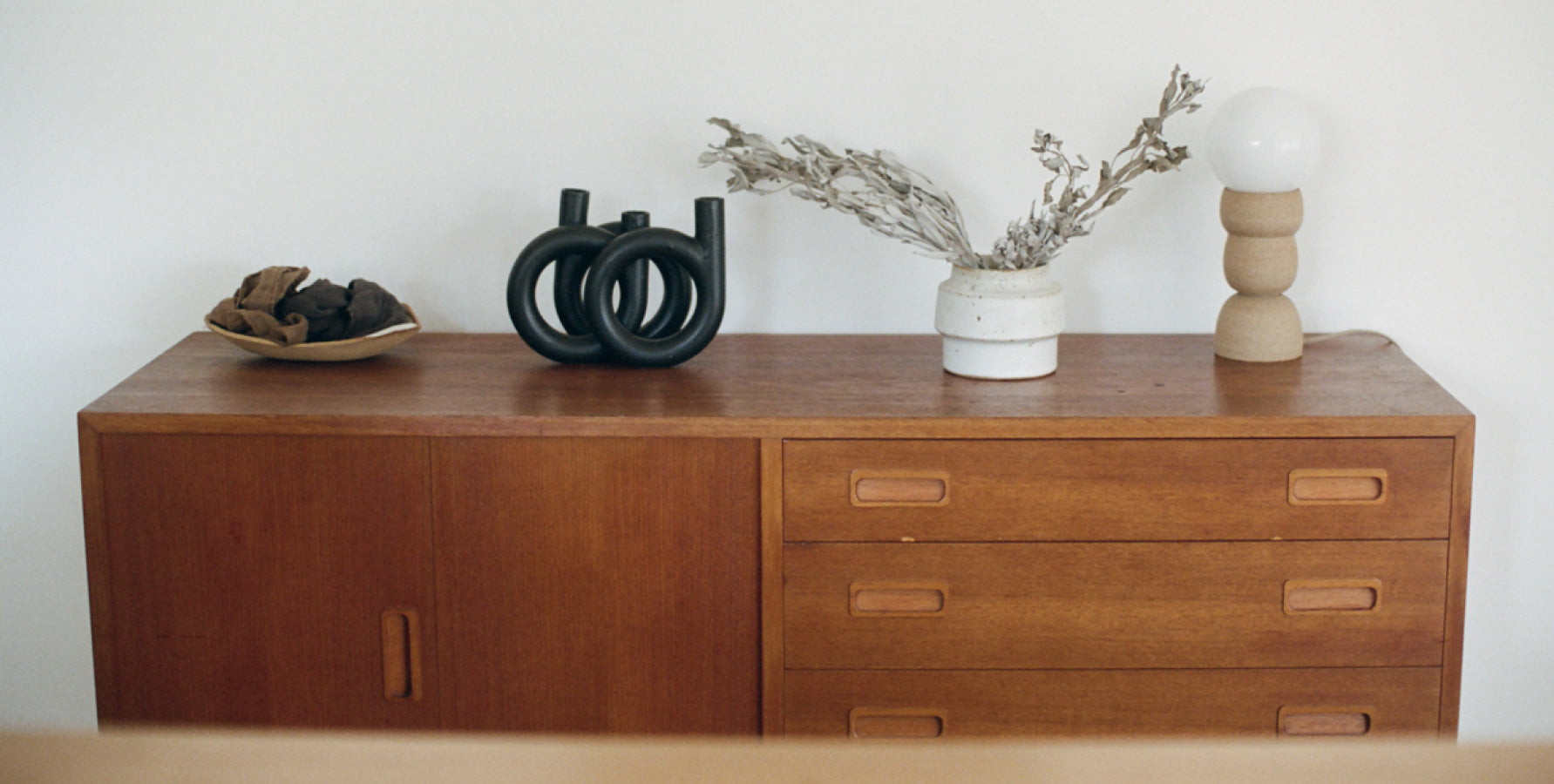 A variety of neutral ceramics sits on top of a vintage wood dresser with drawers against a white wall