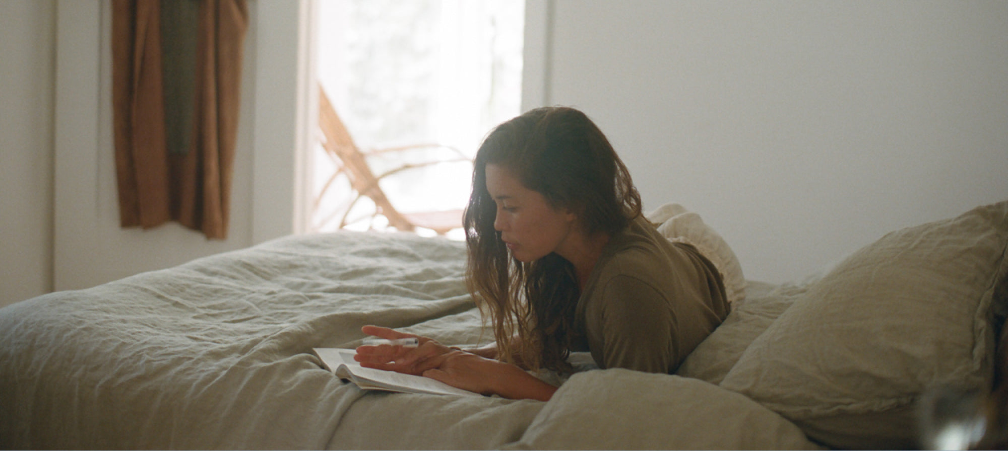Woman laying on a bed writing in a journal on top of sage green linen bedding in a bright bedroom