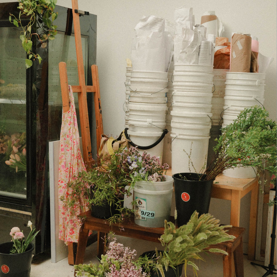 A cluttered workshop filled with stacks of white plastic buckets, potted green plants, floral arrangements, wooden easels, and a glass cabinet against a plain off-white wall.