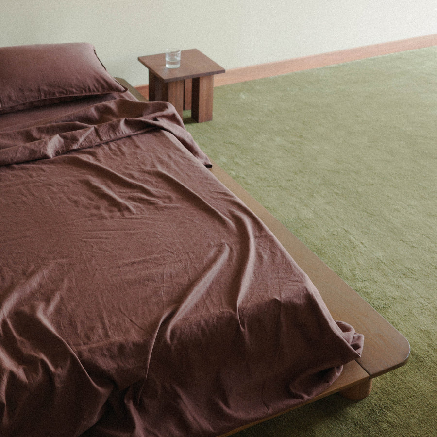 A bed dressed in a rust-brown sheet set with a matching pillow, shown in a minimalist bedroom with a light wood platform bed frame, a small wooden side table, and a sage green rug.