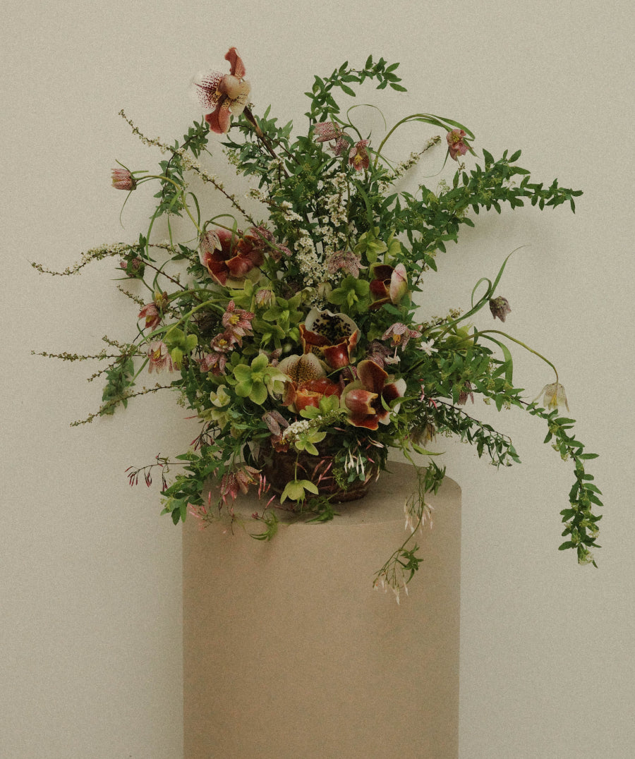 An elaborate floral arrangement featuring green foliage, small white flowers, and reddish-brown blossoms, displayed on a cylindrical beige pedestal against a plain beige wall.