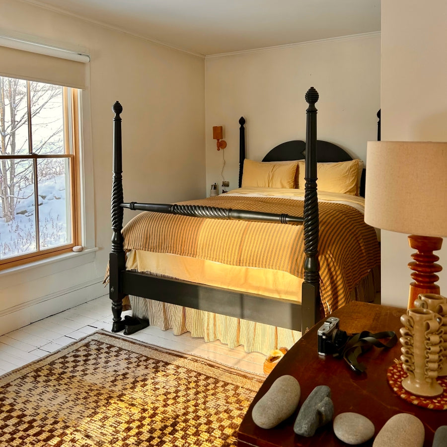 A cozy bedroom scene featuring a black wooden four-poster bed dressed in warm yellow and gold striped bedding, set against cream walls with a window showing a snowy outdoor view.