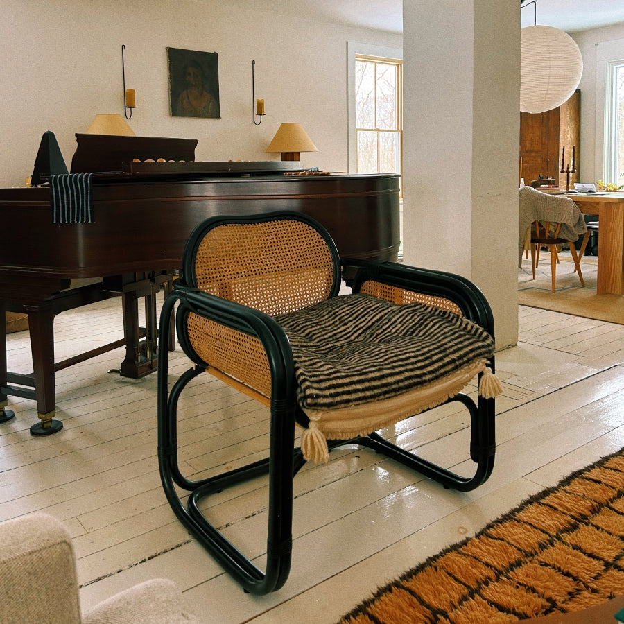 A classic cantilever armchair with a black tubular frame, woven cane backrest, and a striped brown and beige cushion, set in a bright, modern living room next to a dark wood grand piano.