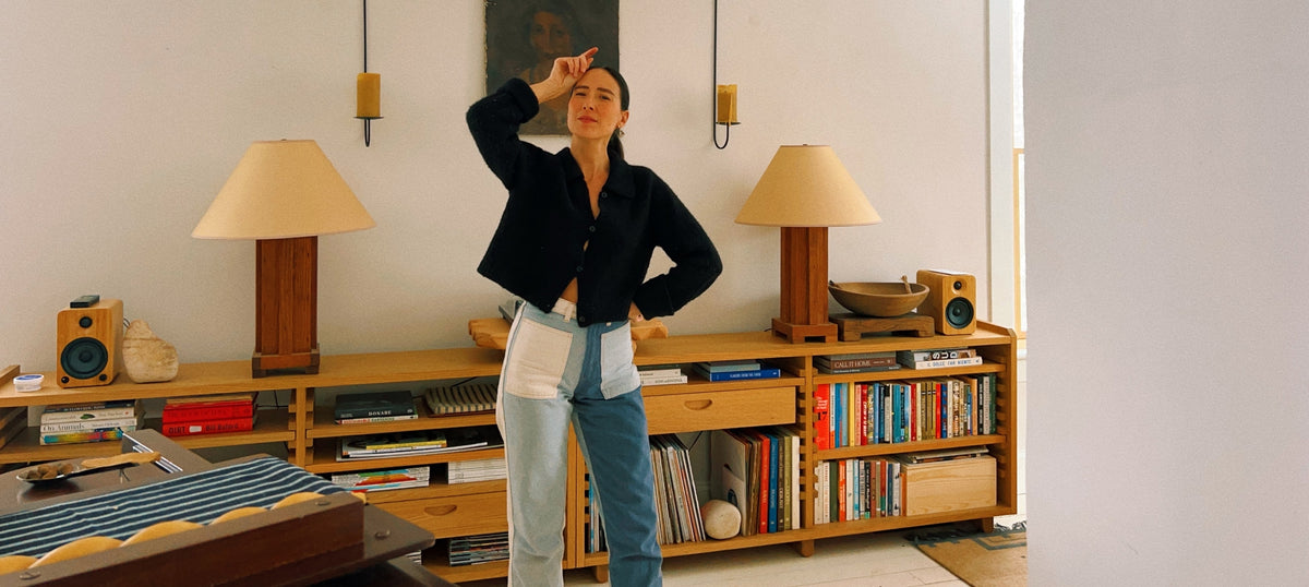 A woman stands in a modern, sunlit room with a wooden bookshelf behind her. She is posing with one hand on her head and the other on her hip.