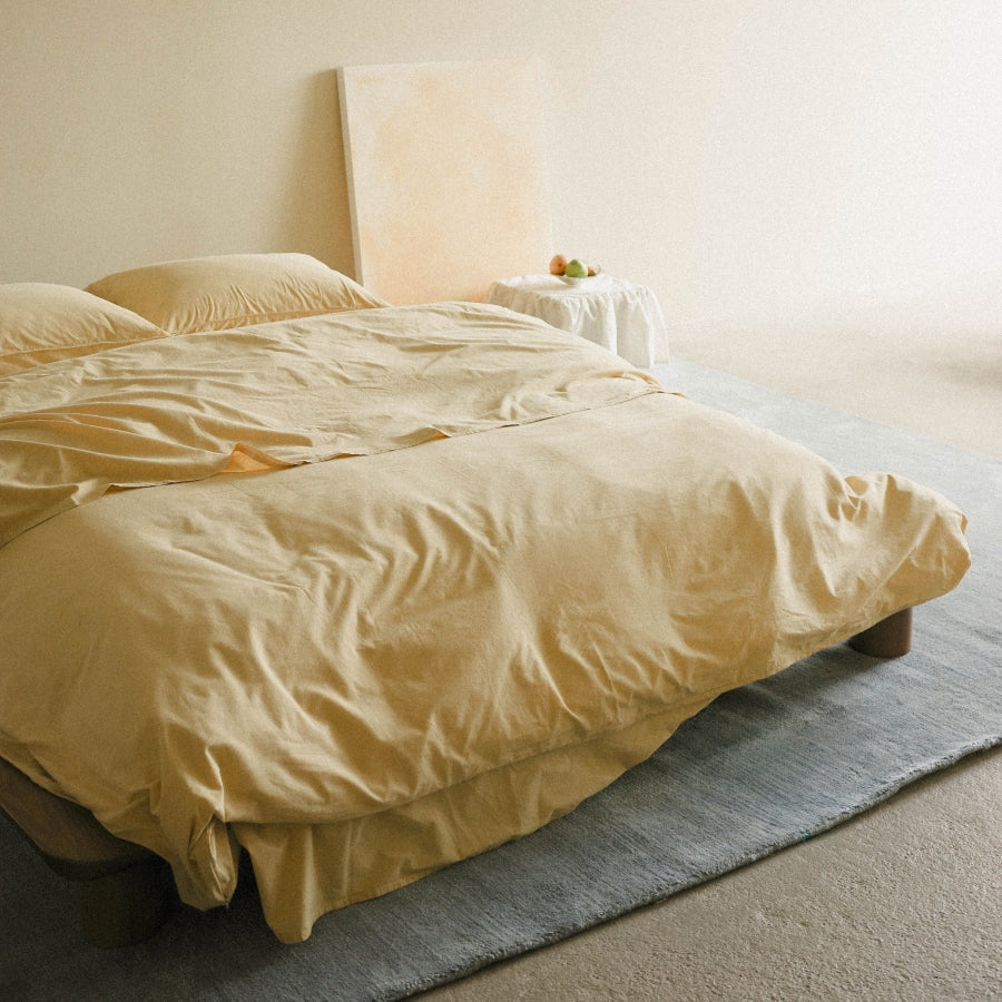 A bed dressed in light yellow bedding, placed in a minimal bedroom with a blue rug, cream walls, and a small side table holding a vase with flowers.