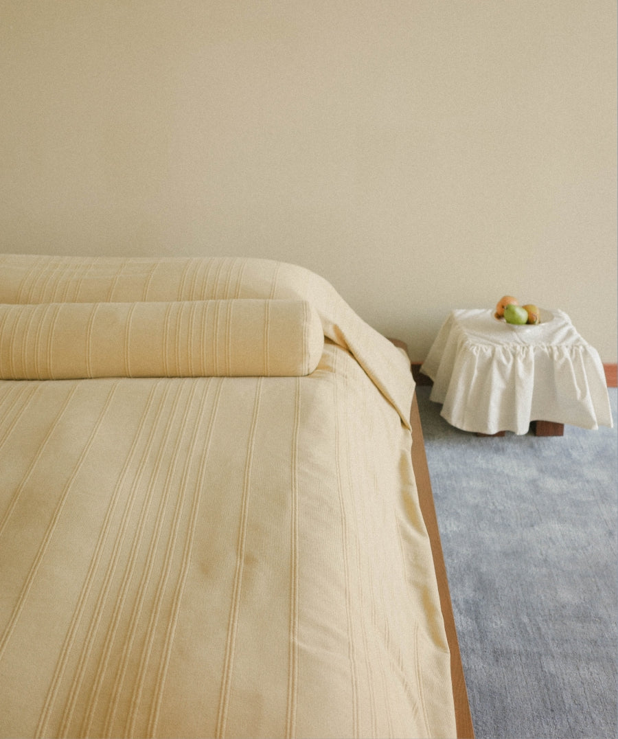 A light yellow bedspread with matching bolster pillow featuring subtle vertical stripes, styled on a bed in a minimalist bedroom setting with a small white-covered side table and a blue rug.