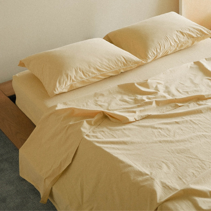 A neatly made bed with light yellow bedding, including two pillows and a rumpled sheet, shown in a minimalist bedroom setting.