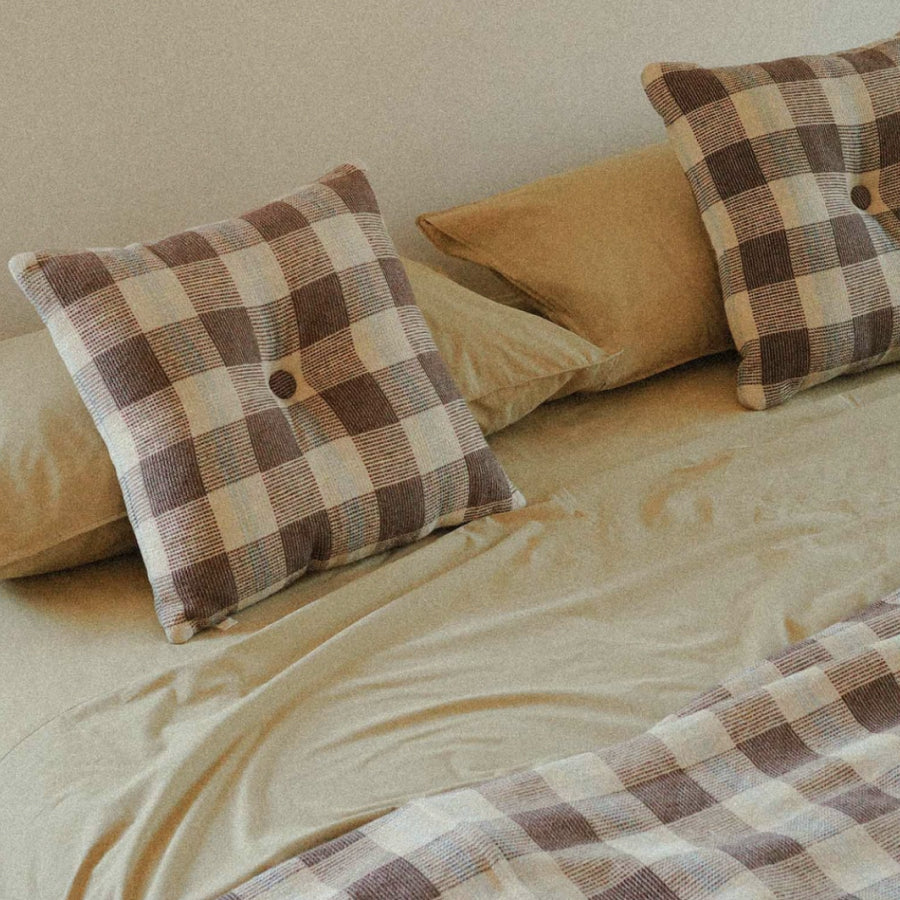Two square throw pillows in a brown, beige, and gray buffalo plaid pattern with button details, styled on a bed with beige sheets and a matching beige pillow.