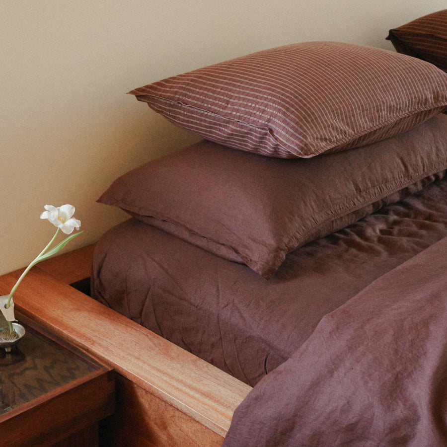 Brown bedding set with matching pillowcases and sheets on a bed, styled in a bedroom setting with a wooden headboard and bedside table holding a small white flower.