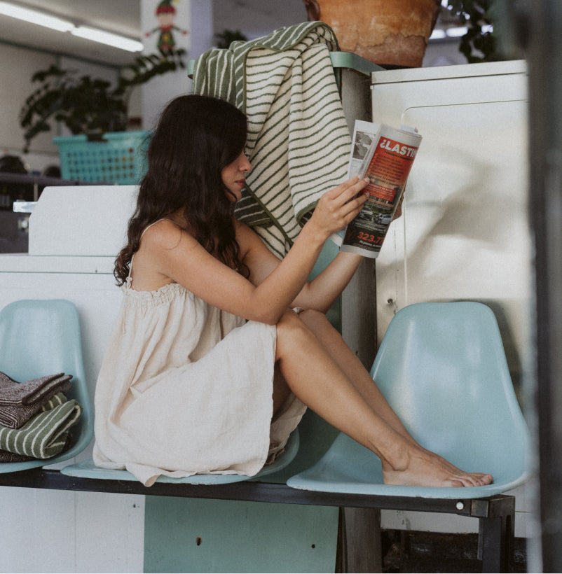 Woman sitting on blue bench seats in a laundromat surrounded by striped bath towels reading a newspaper.
