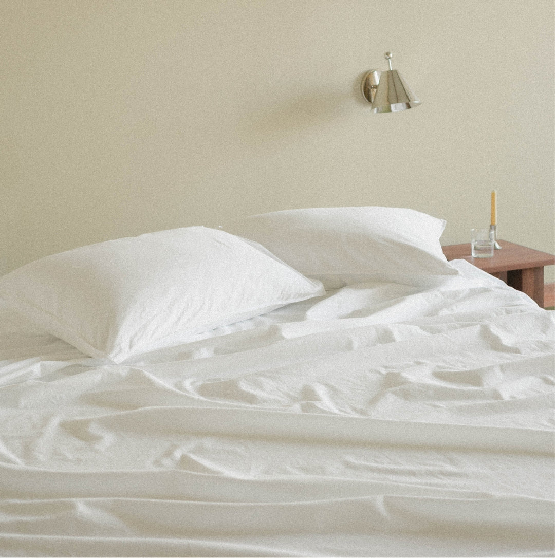 White cotton bed sheets and two white pillows on a bed in a minimalist bedroom with a beige wall, a wall-mounted lamp, and a small wooden side table.