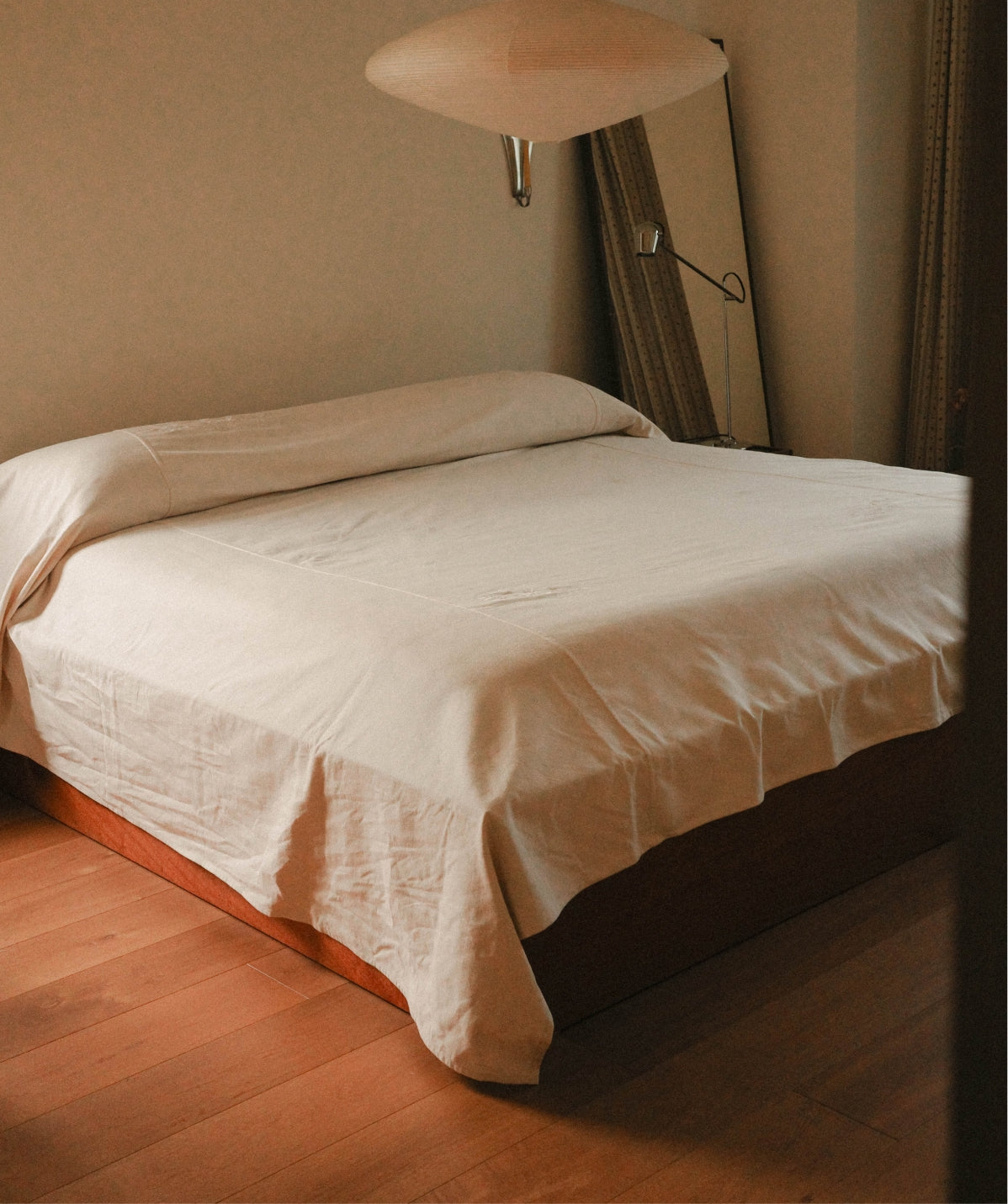 A bed dressed in white linens, positioned in a warmly lit bedroom with wooden flooring, a wall lamp, curtains, and a floor mirror.